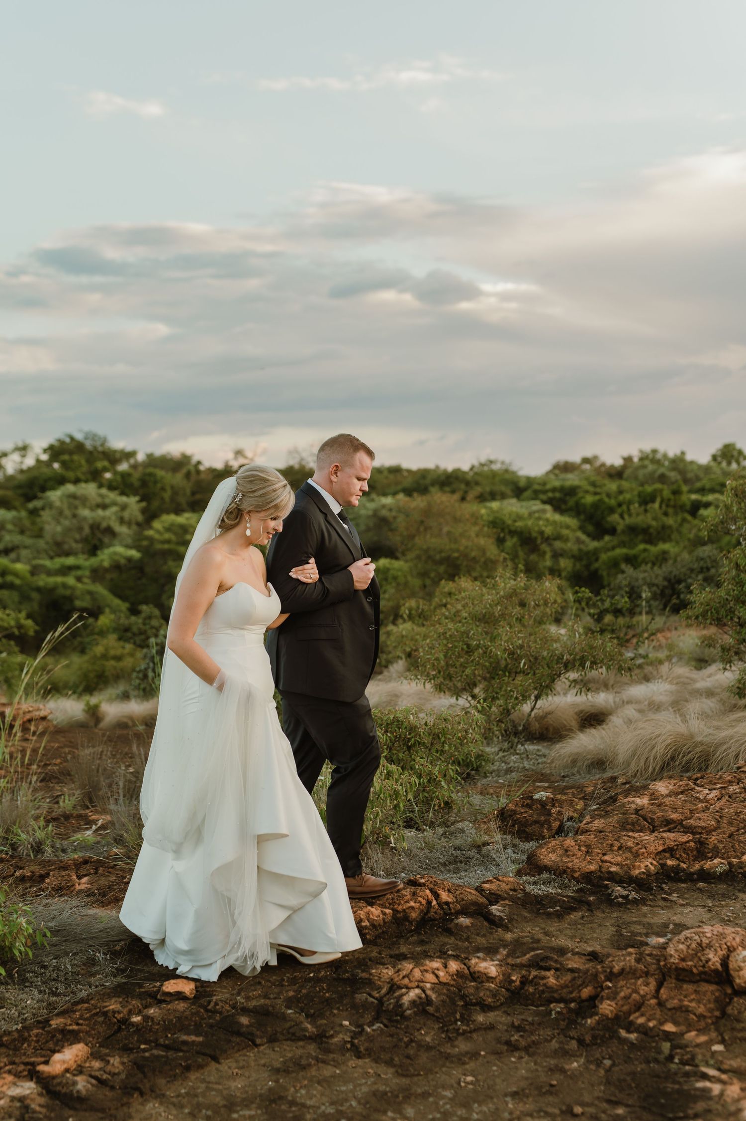 Couple in formal attire walking together on rocky terrain during golden hour.
