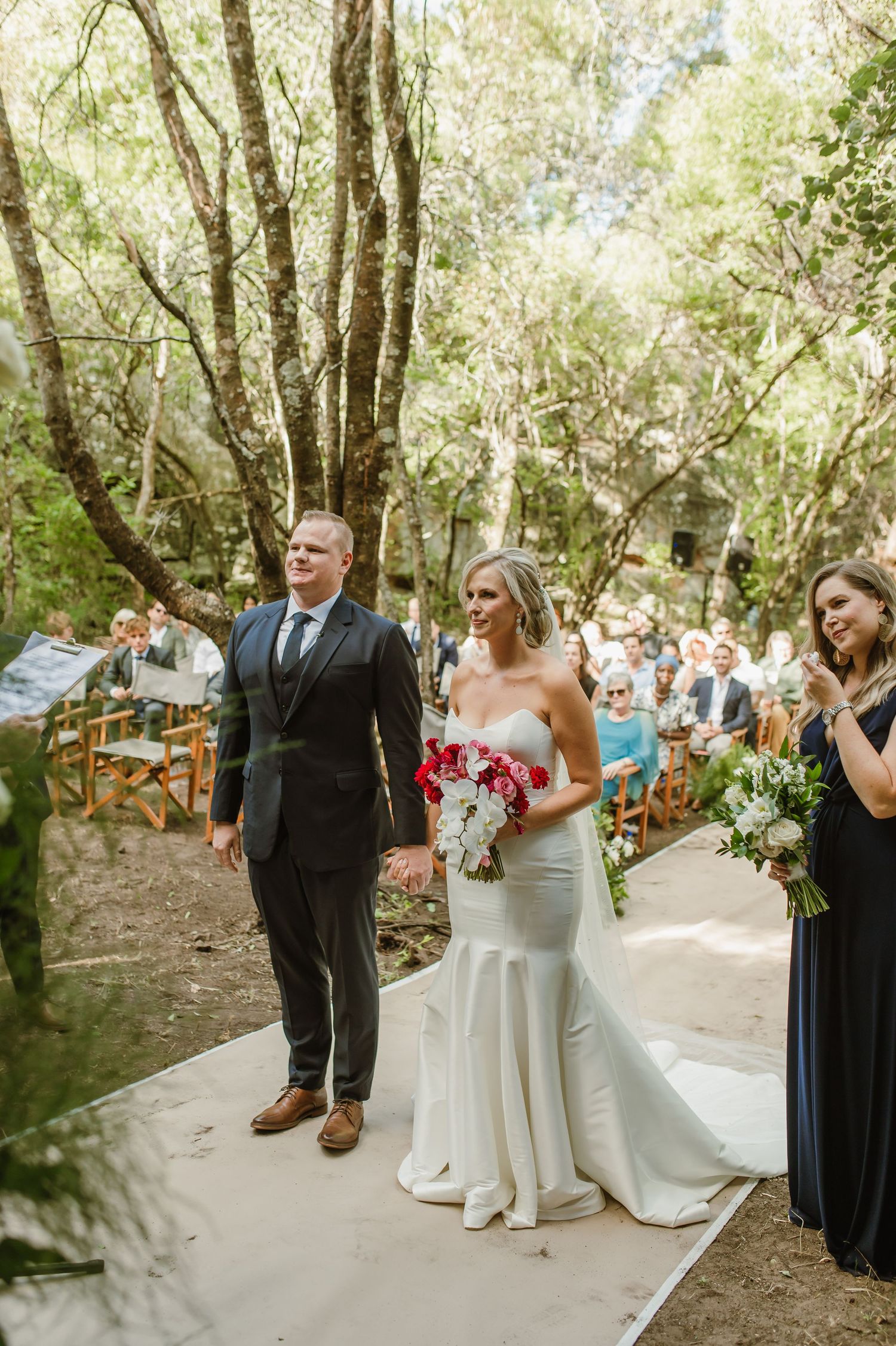 Wedding ceremony participants arranged in line on a forest path with greenery backdrop.