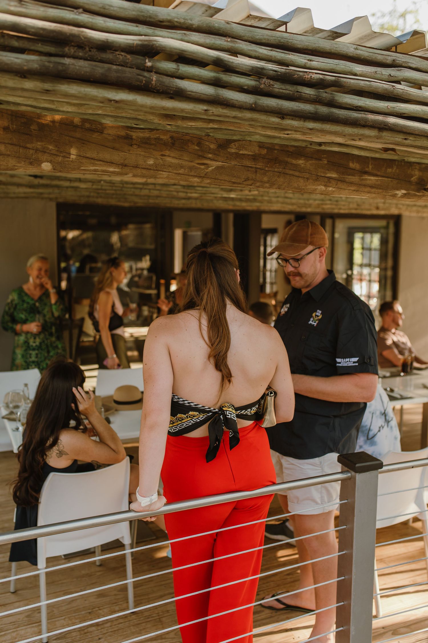 Guests gather at a rustic covered outdoor bar area with wooden beams and metal railings.
