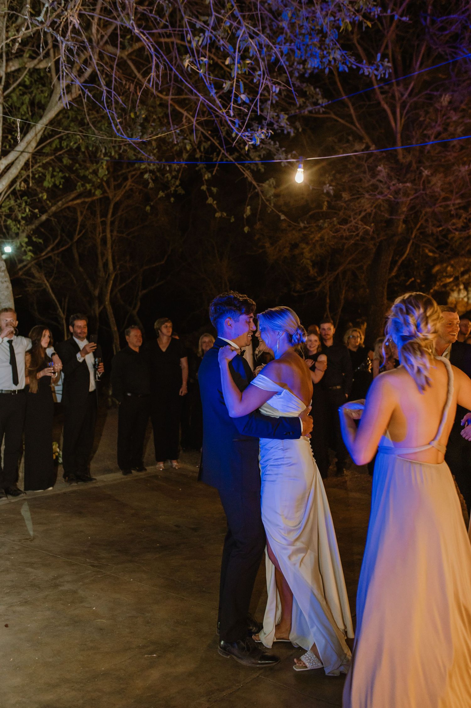 Wedding party dancing together under blue and amber lighting at outdoor reception.
