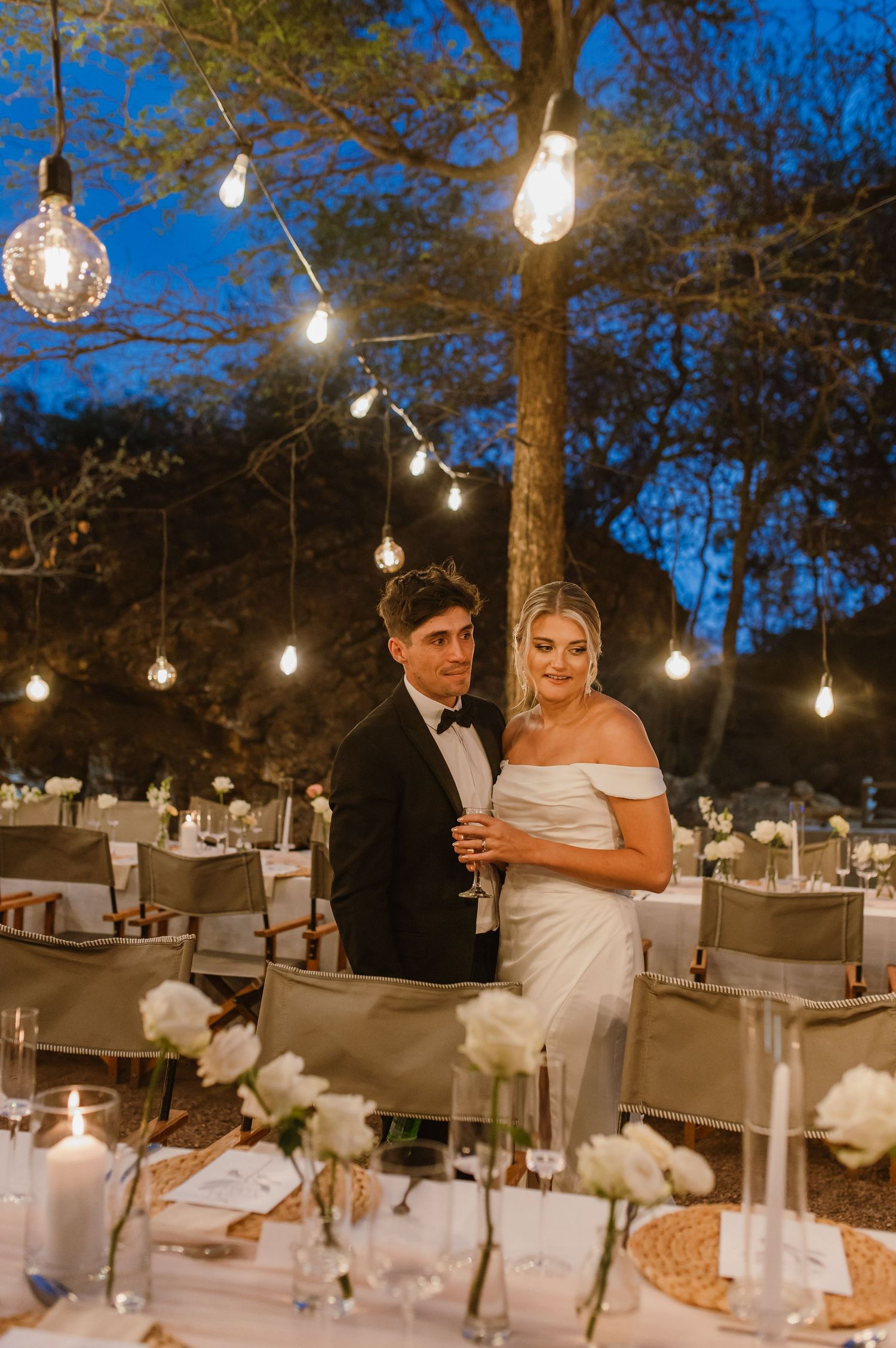 Evening wedding reception tables decorated with white linens and string lights hanging from trees above.