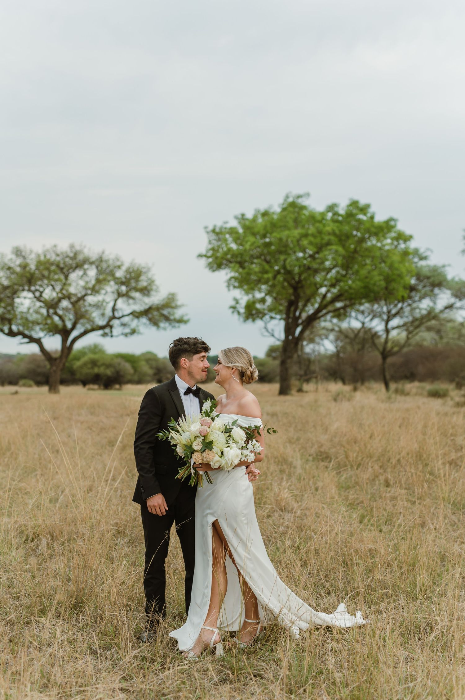 Newlyweds embrace in a sun-lit field with scattered acacia trees in the background.