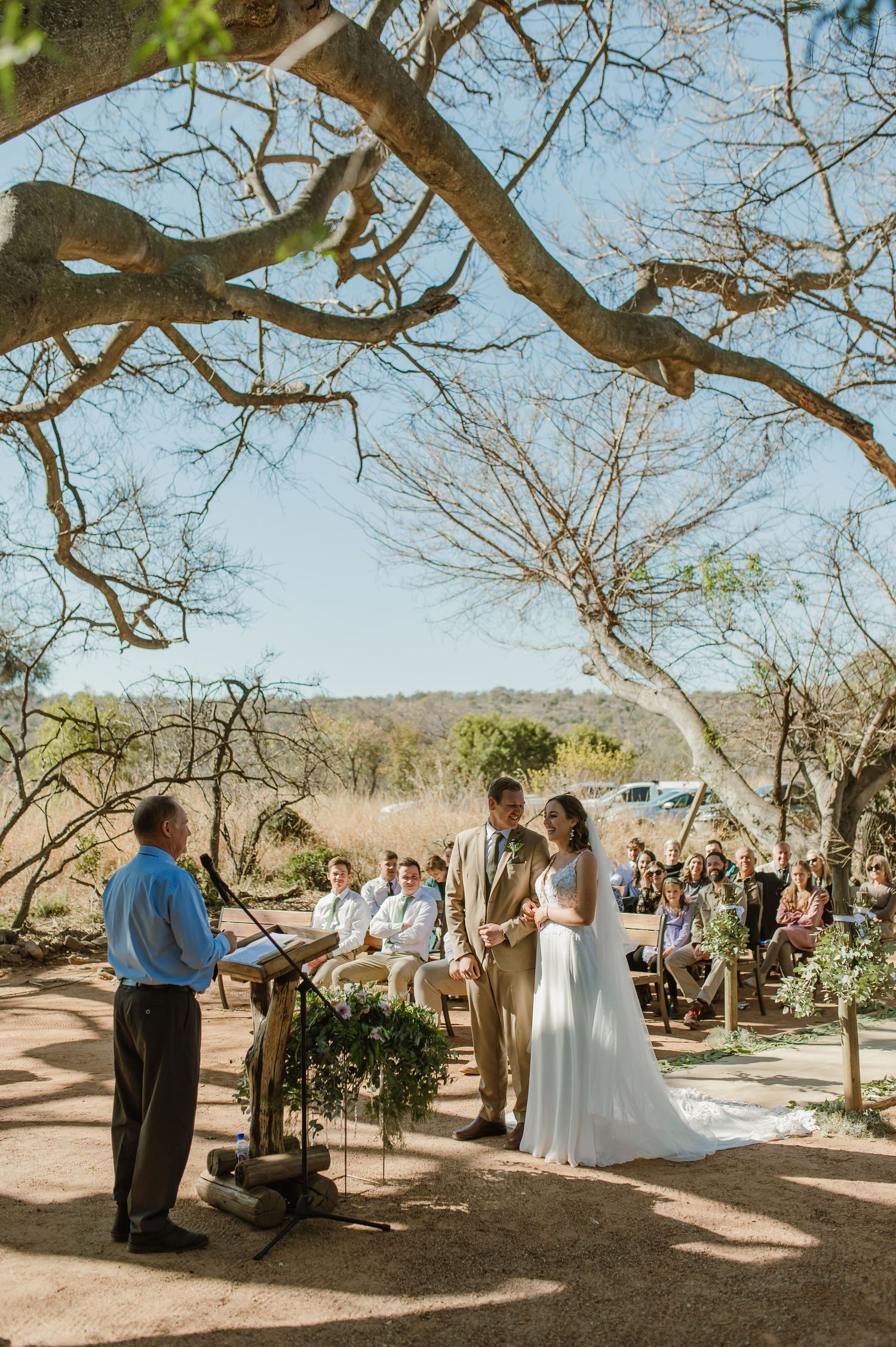 Wedding ceremony taking place beneath sprawling tree branches in a desert landscape with wooden bench seating.
