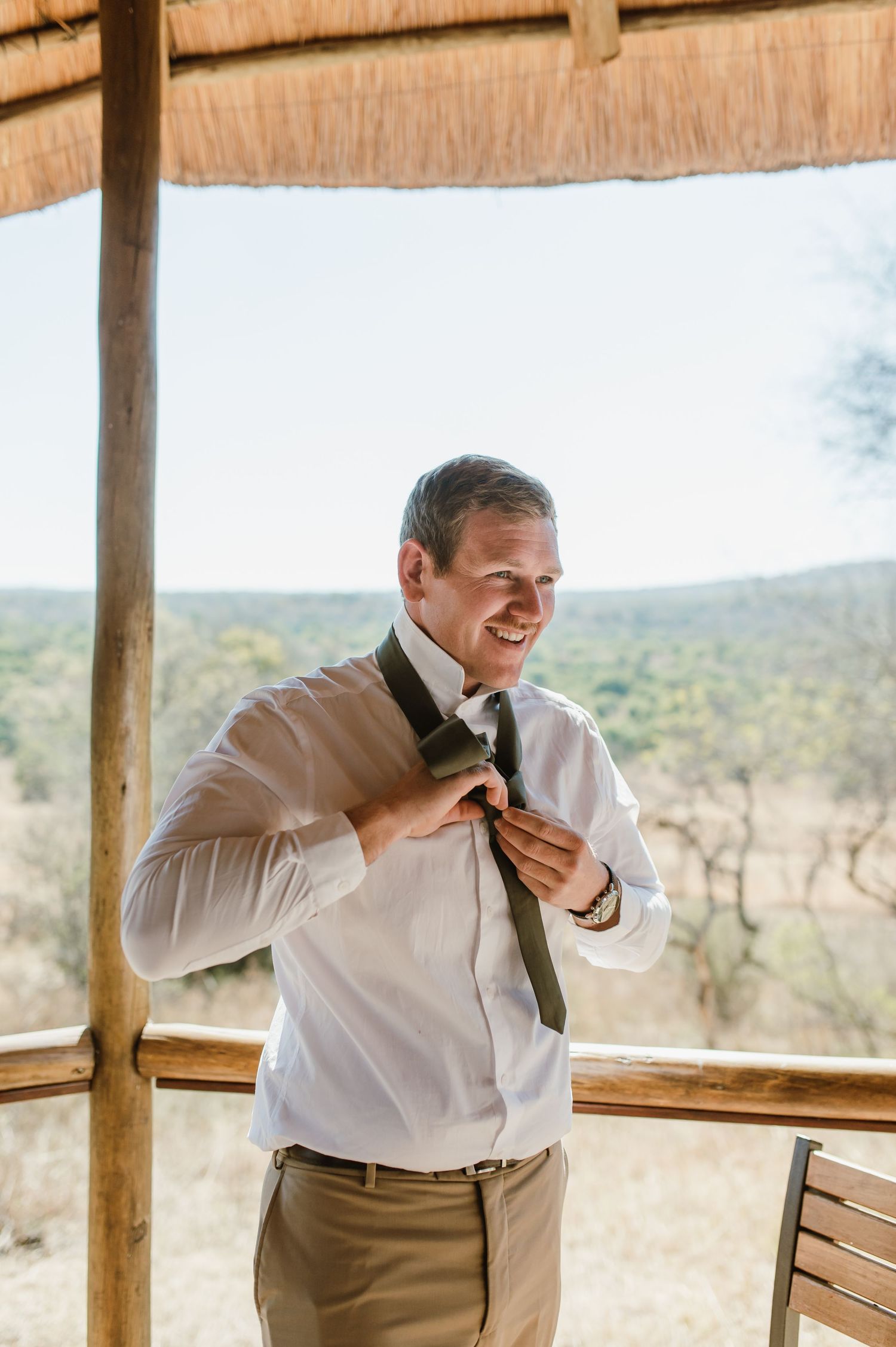 Person in white shirt adjusting tie in thatched-roof gazebo.