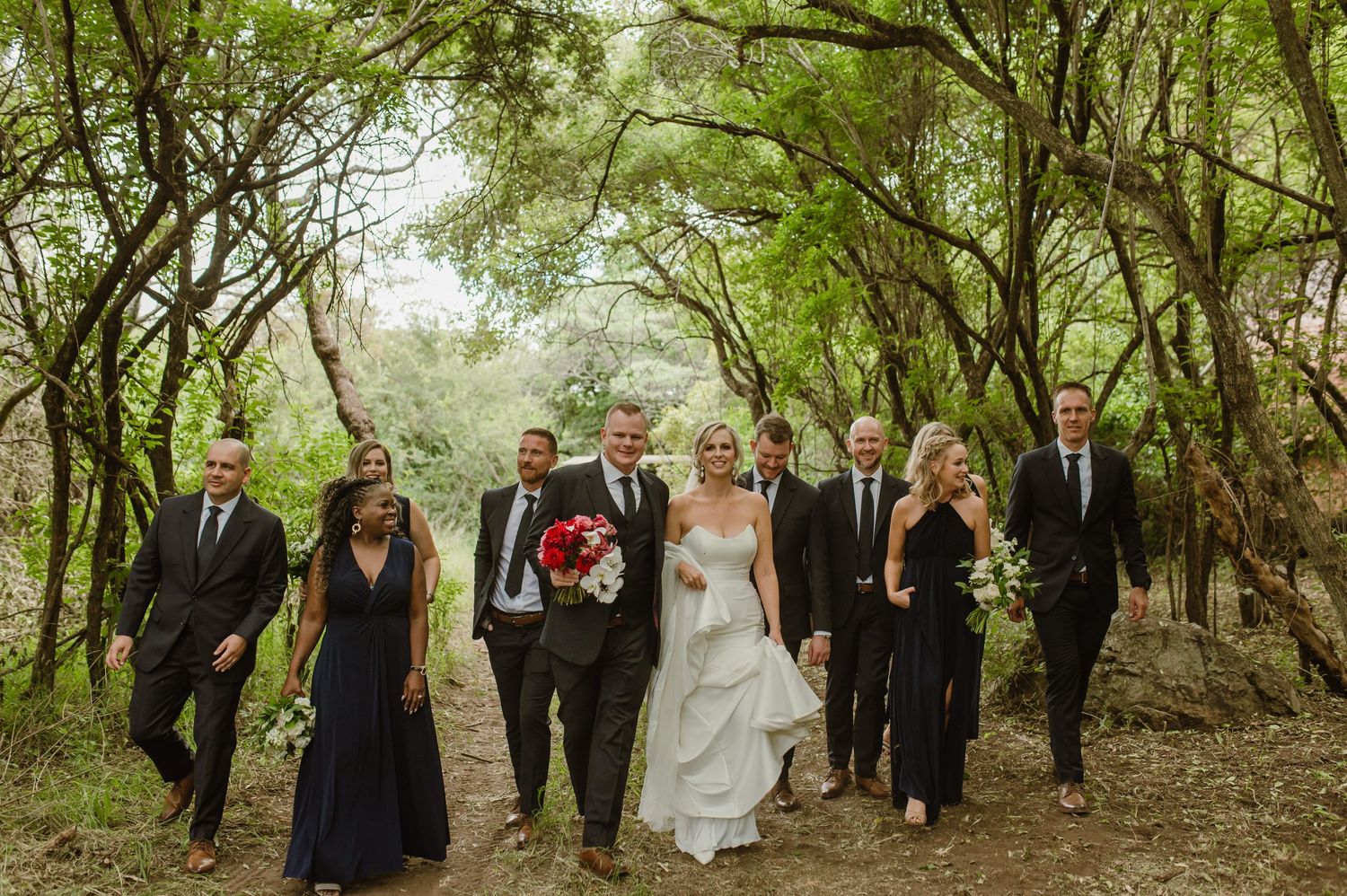 A wedding party walks together through a green wooded area during celebrations.
