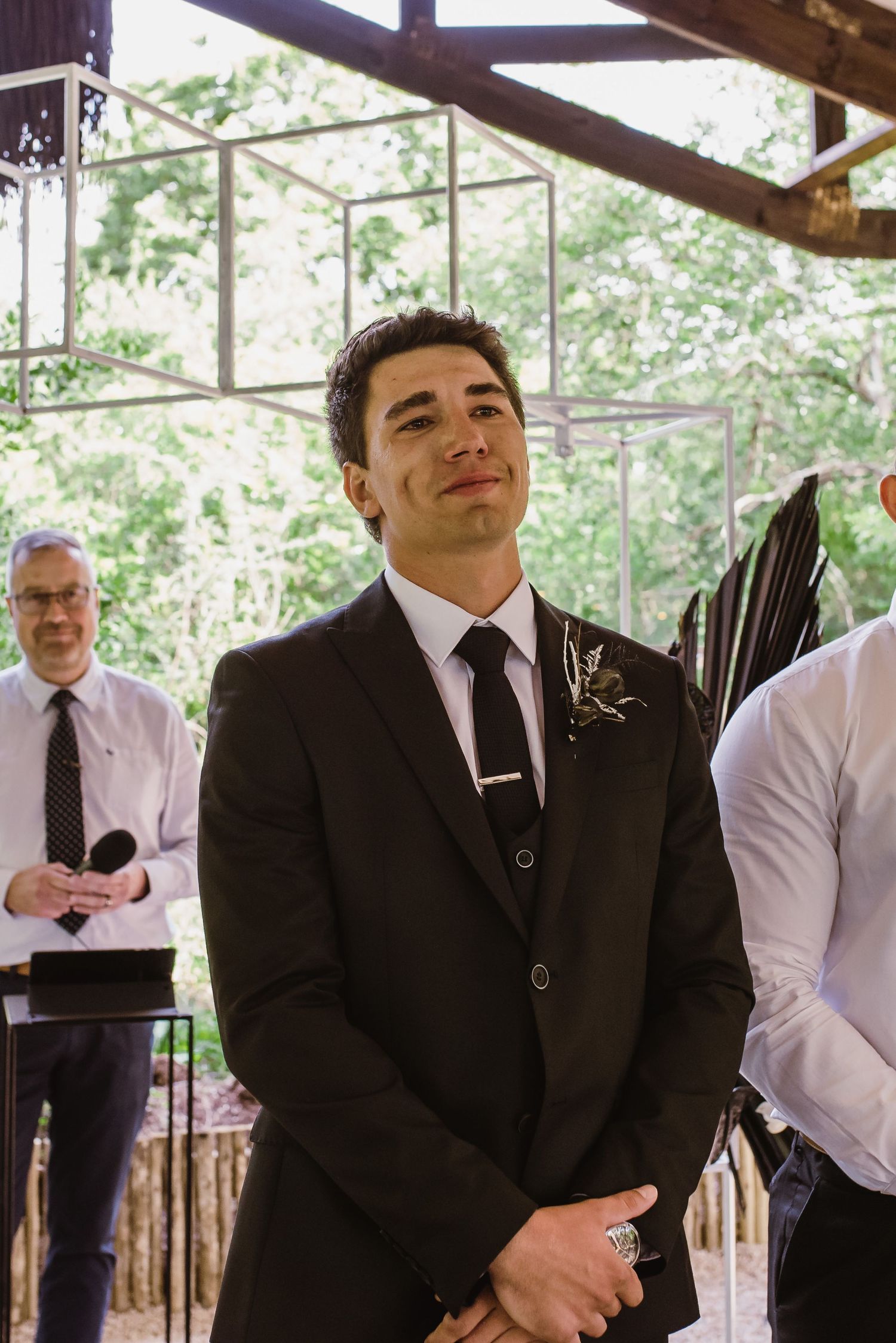 Groomsmen in black suits and white shirts standing in line during outdoor wedding ceremony.