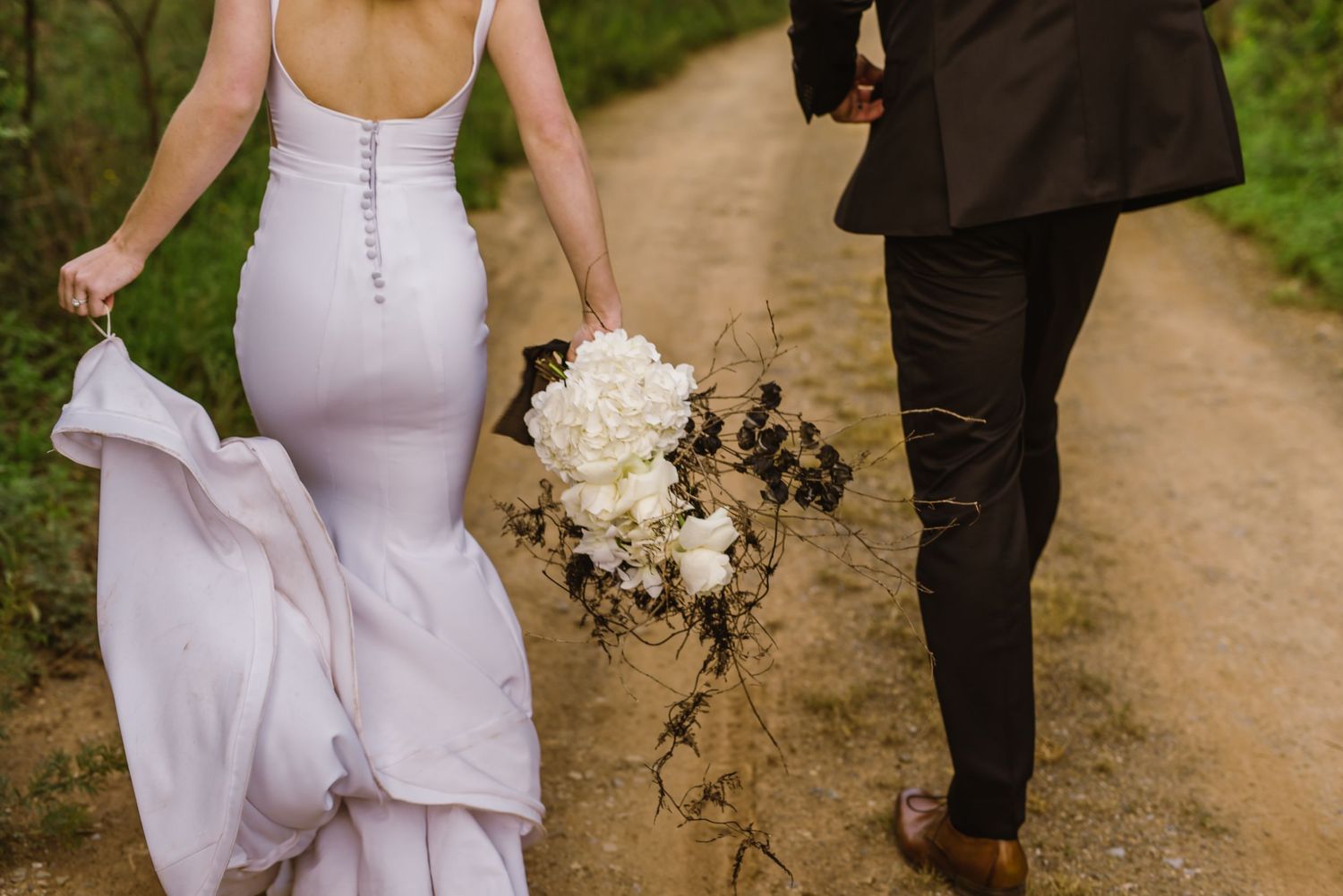 Bride in white dress walks with groom while holding bouquet on desert path.