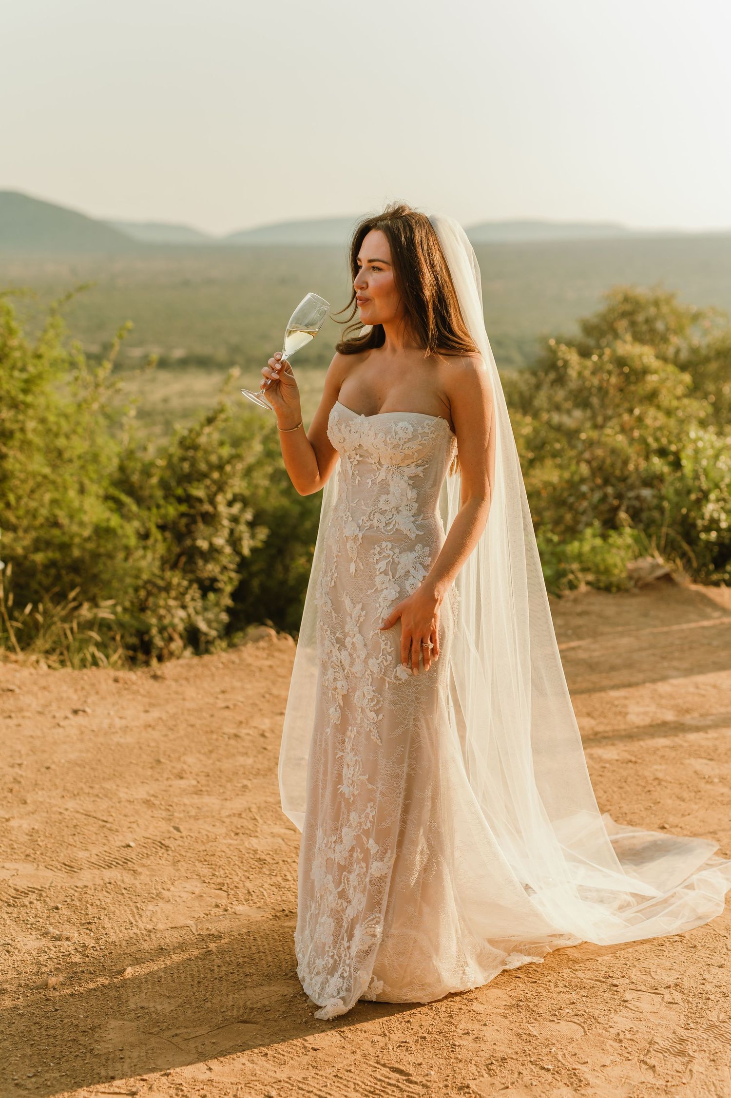 Bride in strapless lace wedding gown and veil sips champagne in scenic outdoor setting.