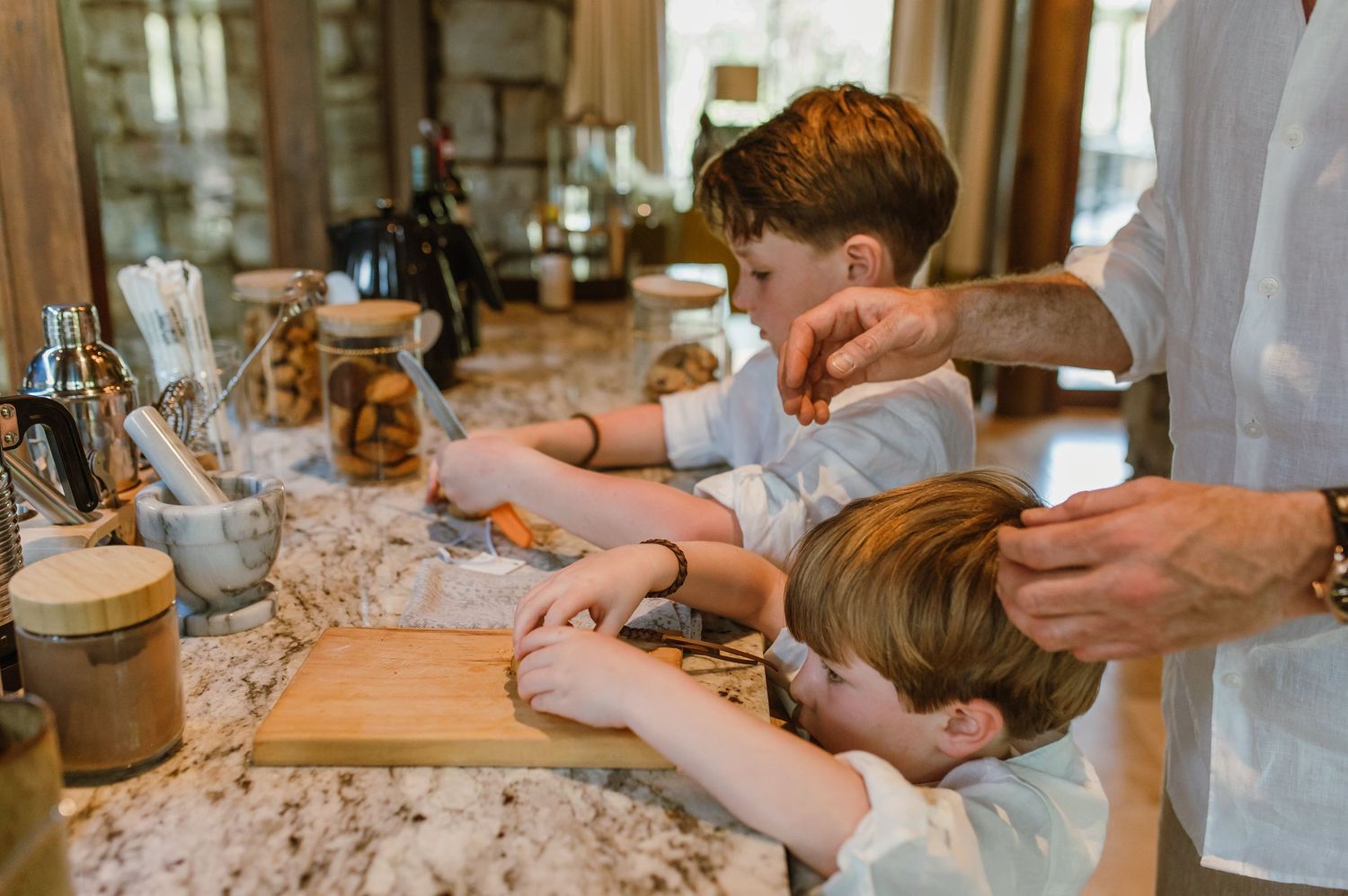 Children and adult preparing food together on a marble kitchen counter with wooden cutting board.