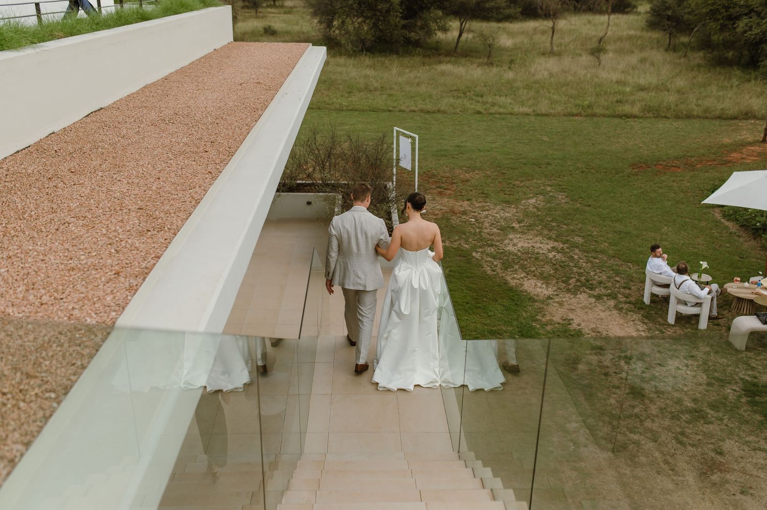 A couple in wedding attire walks down an outdoor path alongside a modern architectural wall with glass panels.