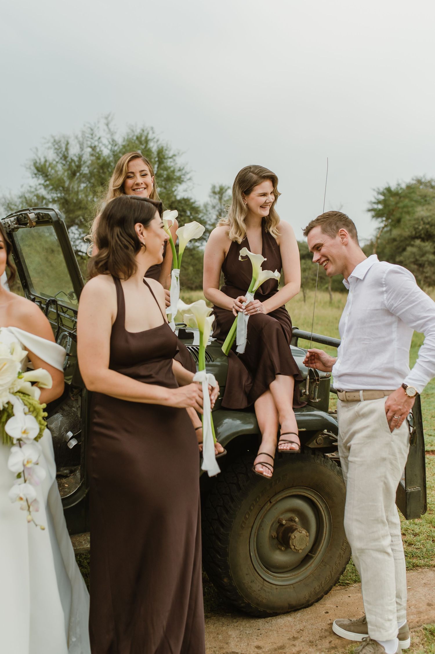 Wedding party in brown dresses and white attire pose playfully near vintage military jeep during outdoor celebration.