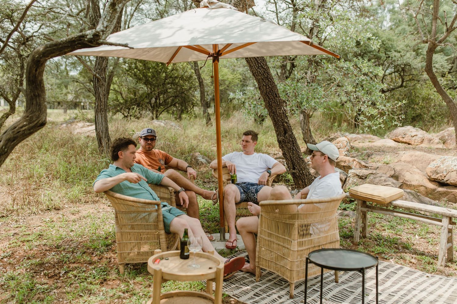 A group relaxes outdoors under a patio umbrella on wicker furniture surrounded by trees in a natural setting.