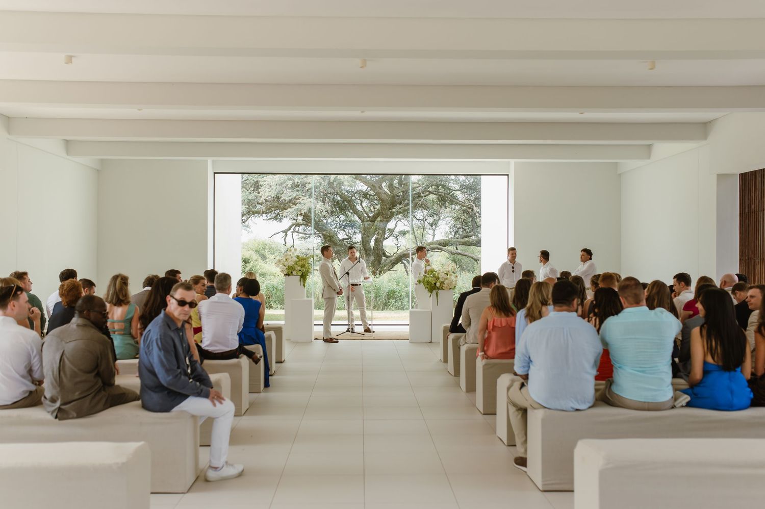 Wedding guests seated in rows at a modern white venue with large windows overlooking trees.