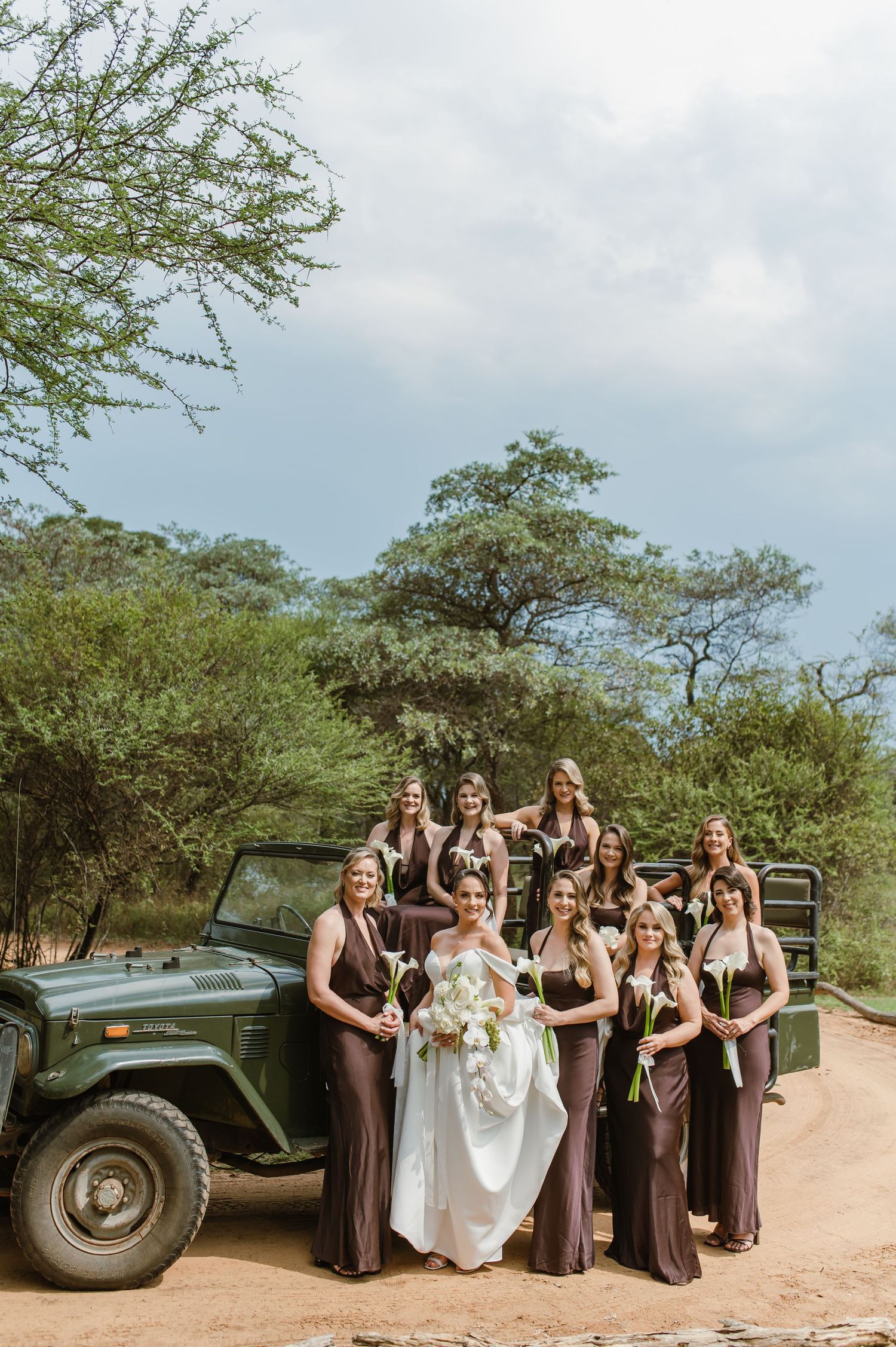 Wedding party posing with vintage military vehicle in tropical outdoor setting.