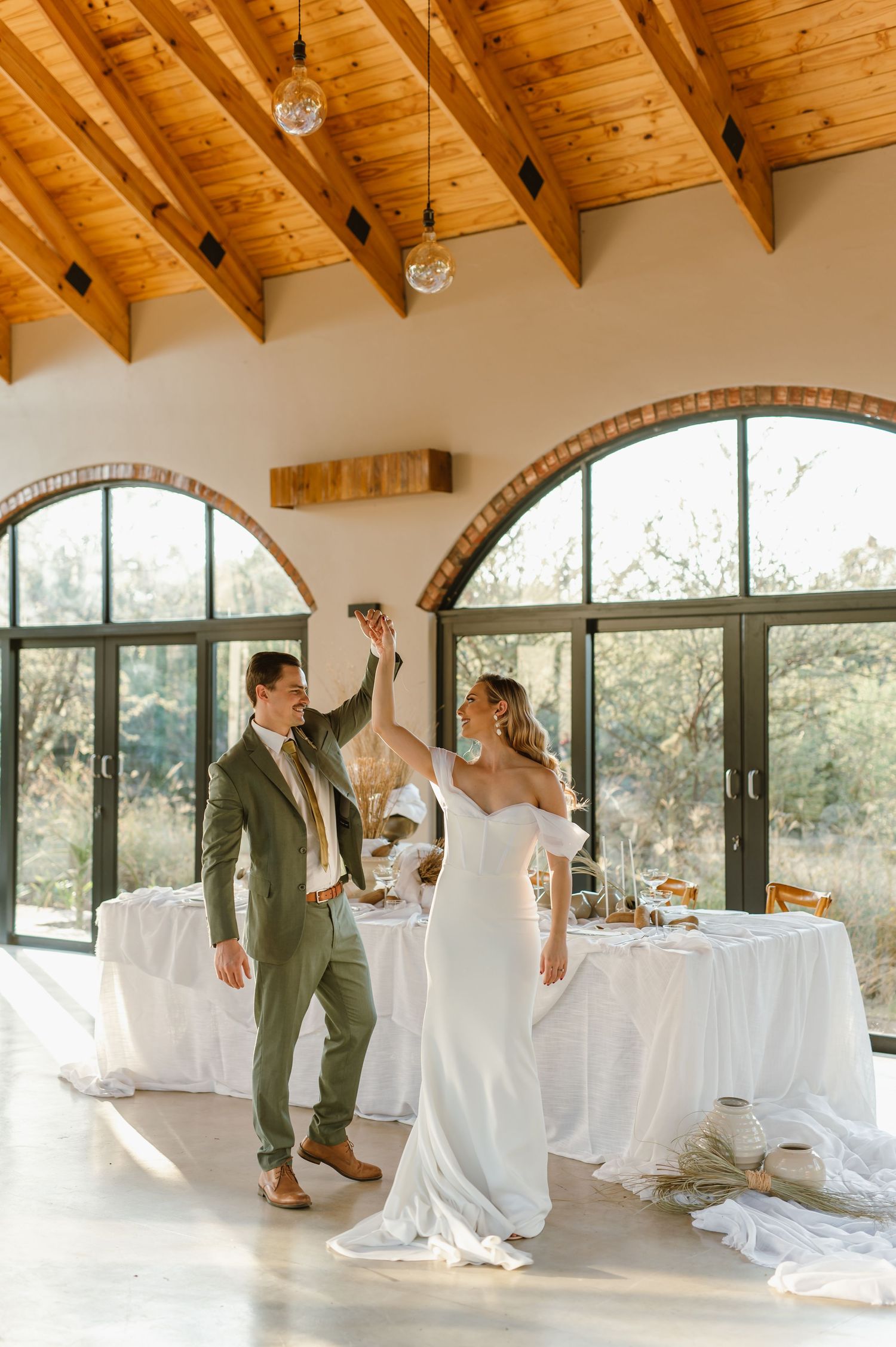 Newlyweds dancing in sunlit venue with wooden beams and arched windows.