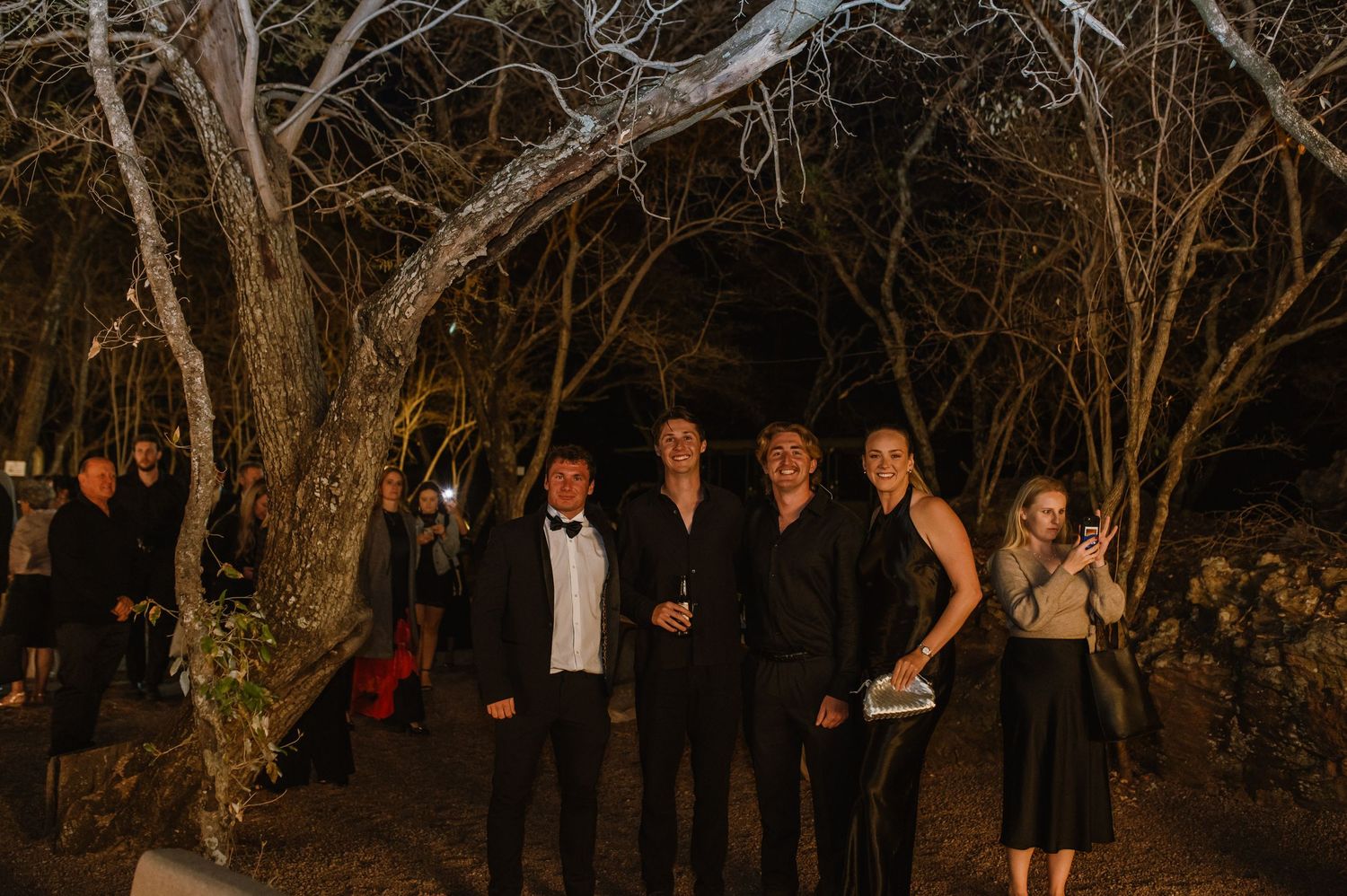 Wedding guests mingle under string lights and trees at an outdoor evening reception venue.