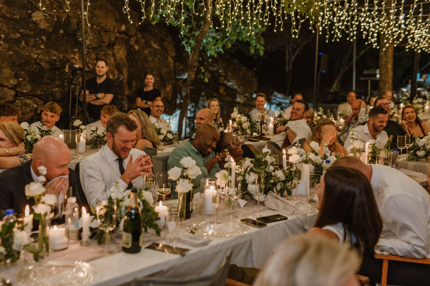 Wedding reception dinner under twinkling fairy lights with elegant table settings and floral centerpieces.