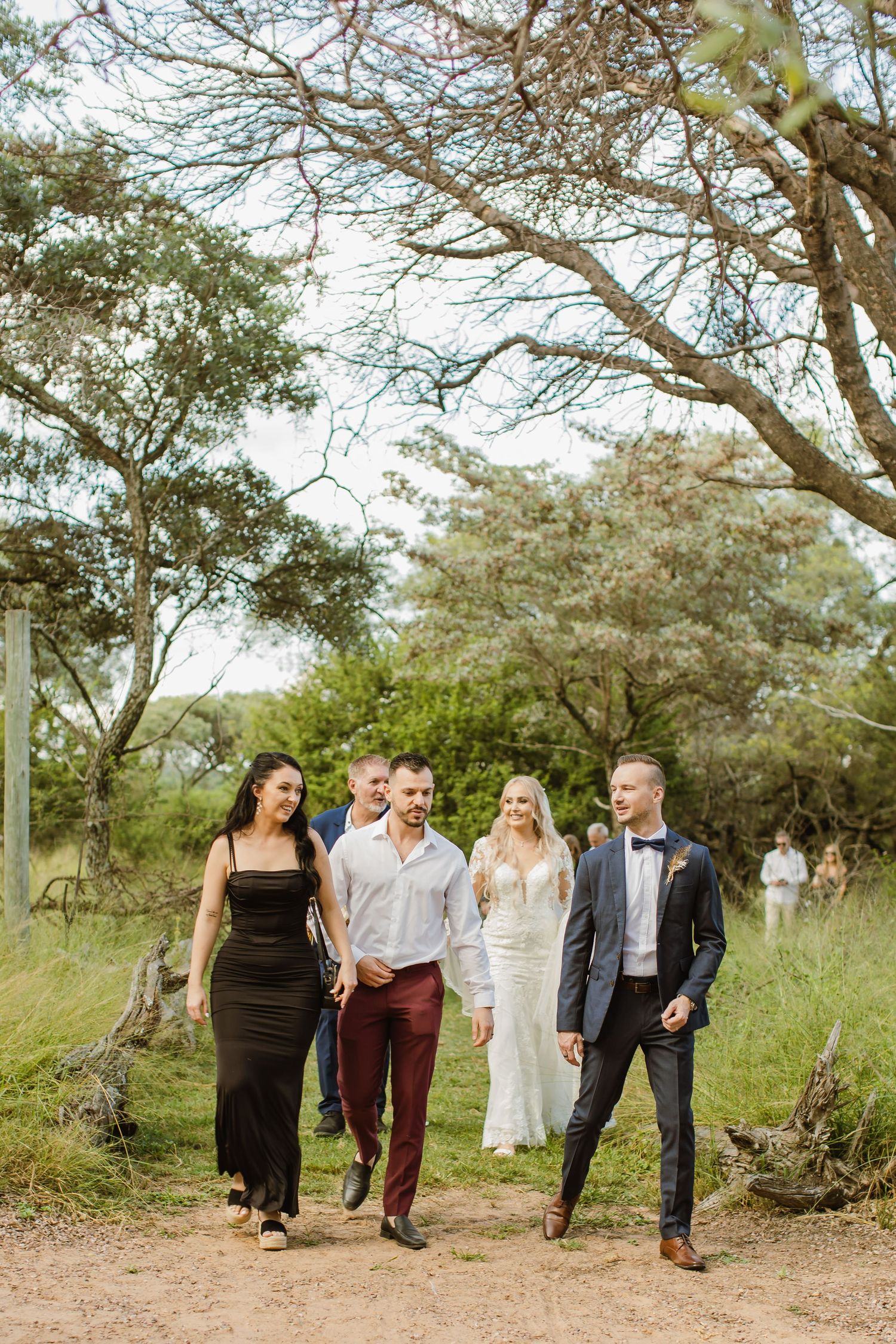 Group photo of wedding party members walking together on a dirt path surrounded by trees.