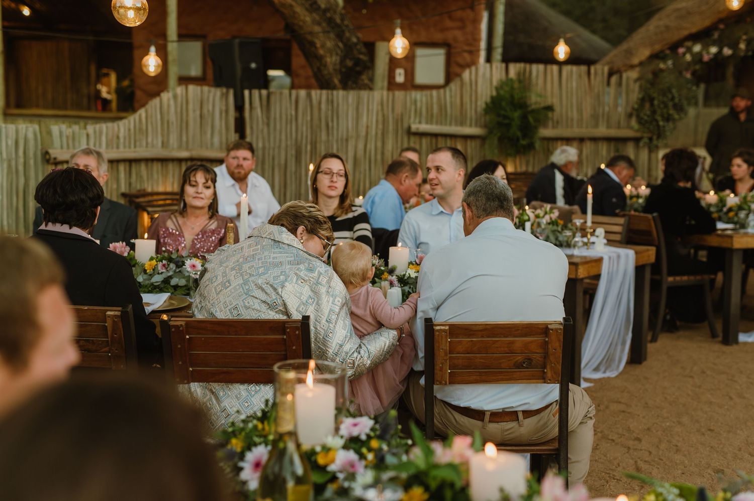 Evening wedding reception with hanging lights and long wooden tables in an outdoor setting.