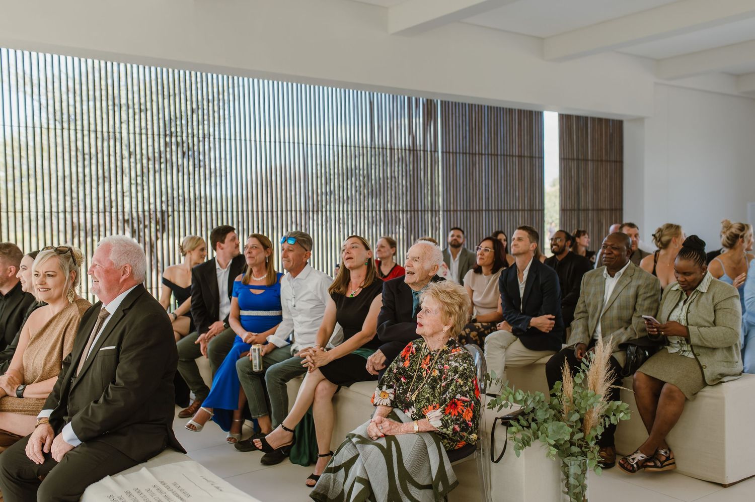 Wedding guests seated in rows at an indoor ceremony with large windows and natural light.