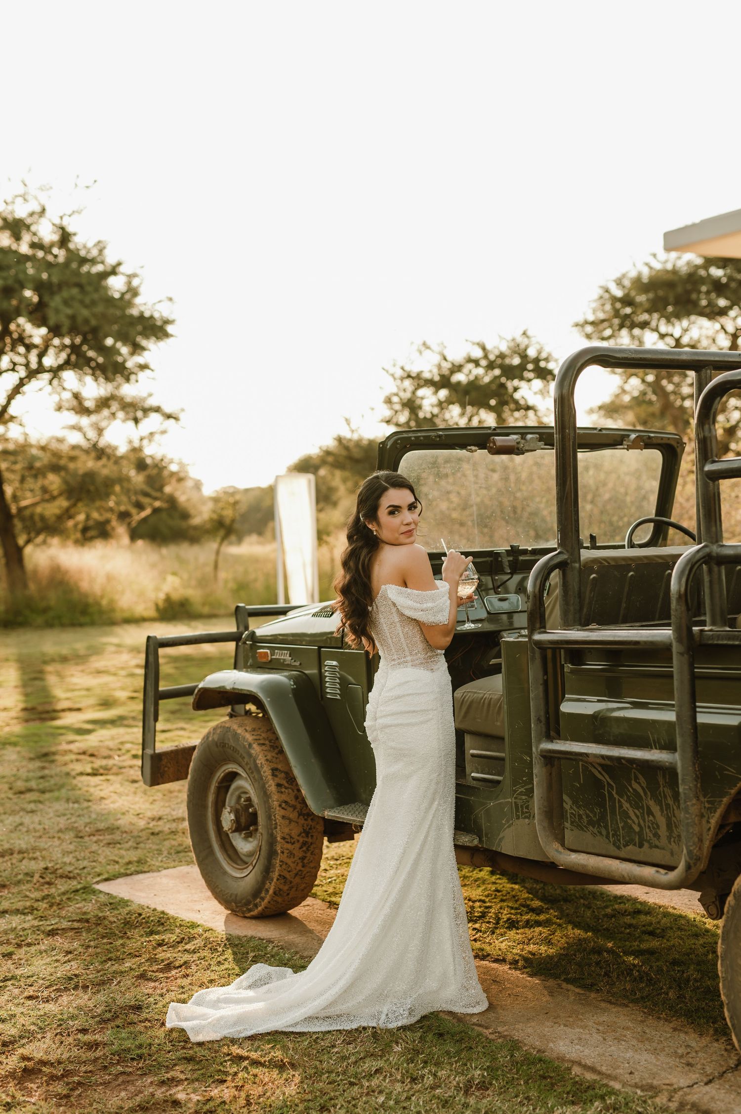 Bride in white dress poses with vintage off-road vehicle during golden hour.