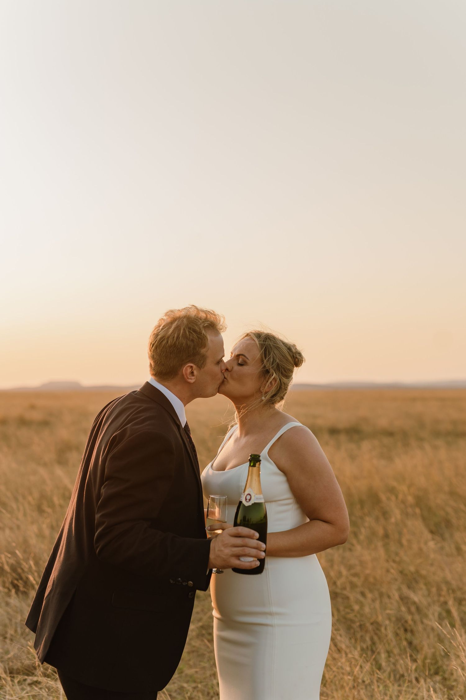Romantic couple sharing champagne and kisses in a sunlit field.