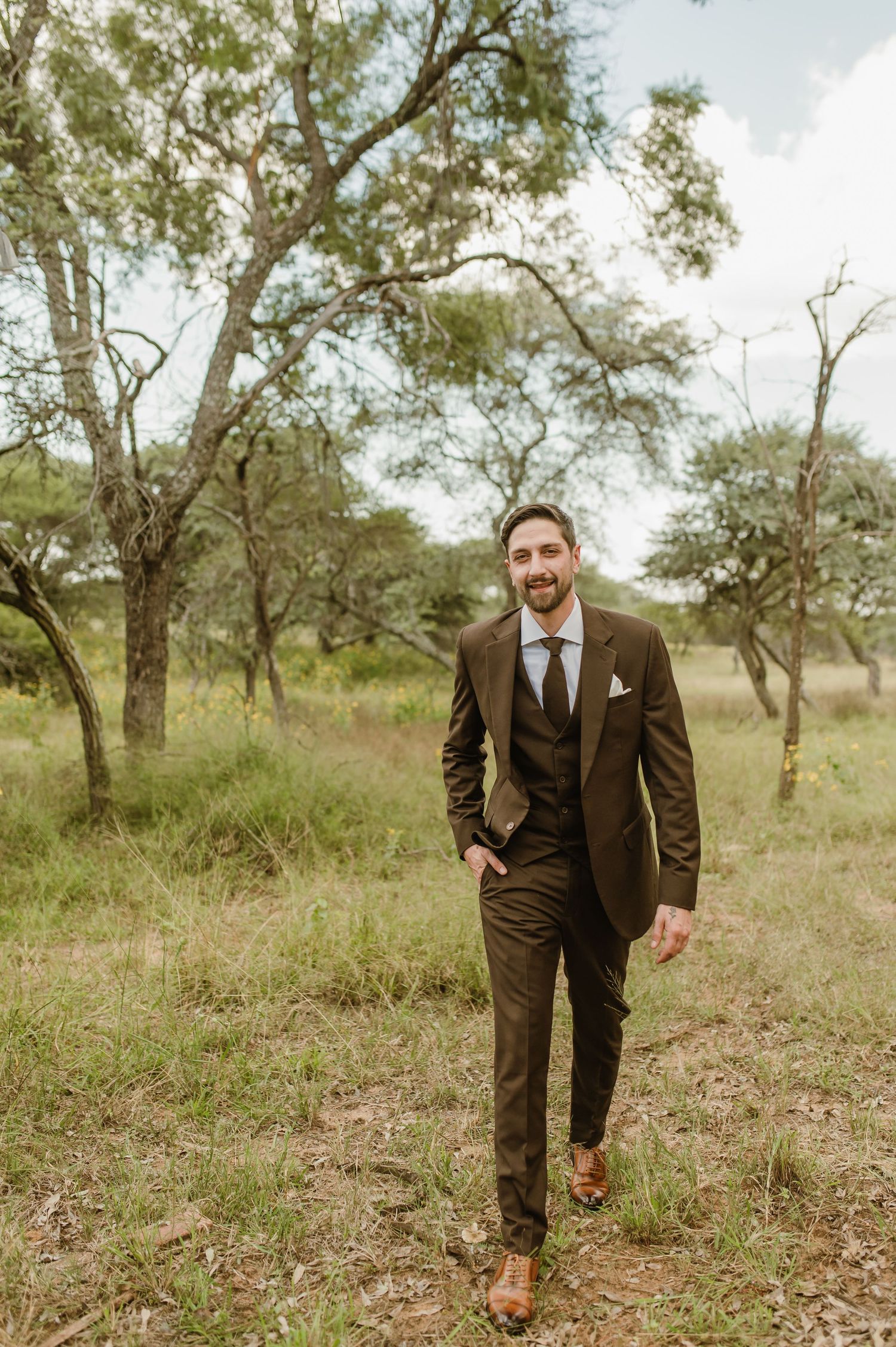 Person in brown three-piece suit walking through natural woodland setting.