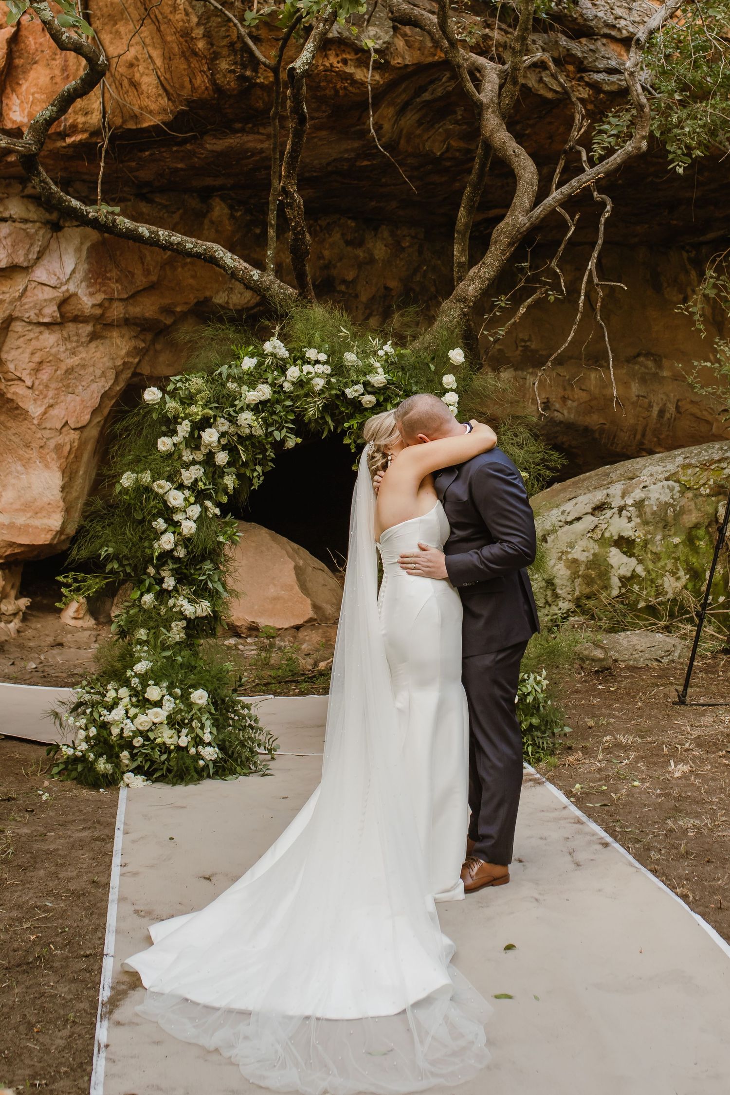 A romantic wedding first look moment in front of a floral arch against a rocky cliff backdrop.