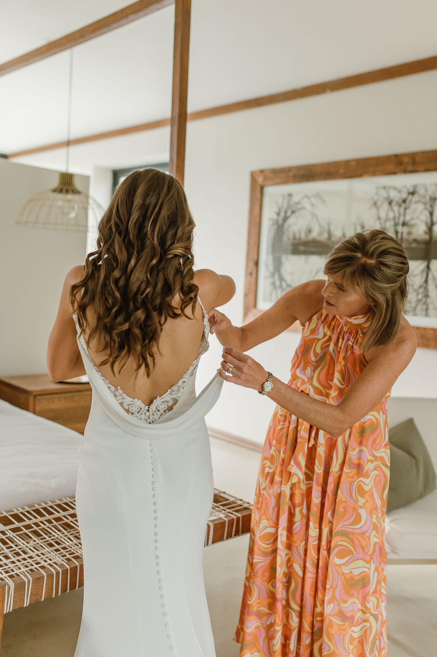 A wedding dress being fastened by someone in a peach floral dress in a sunlit room.