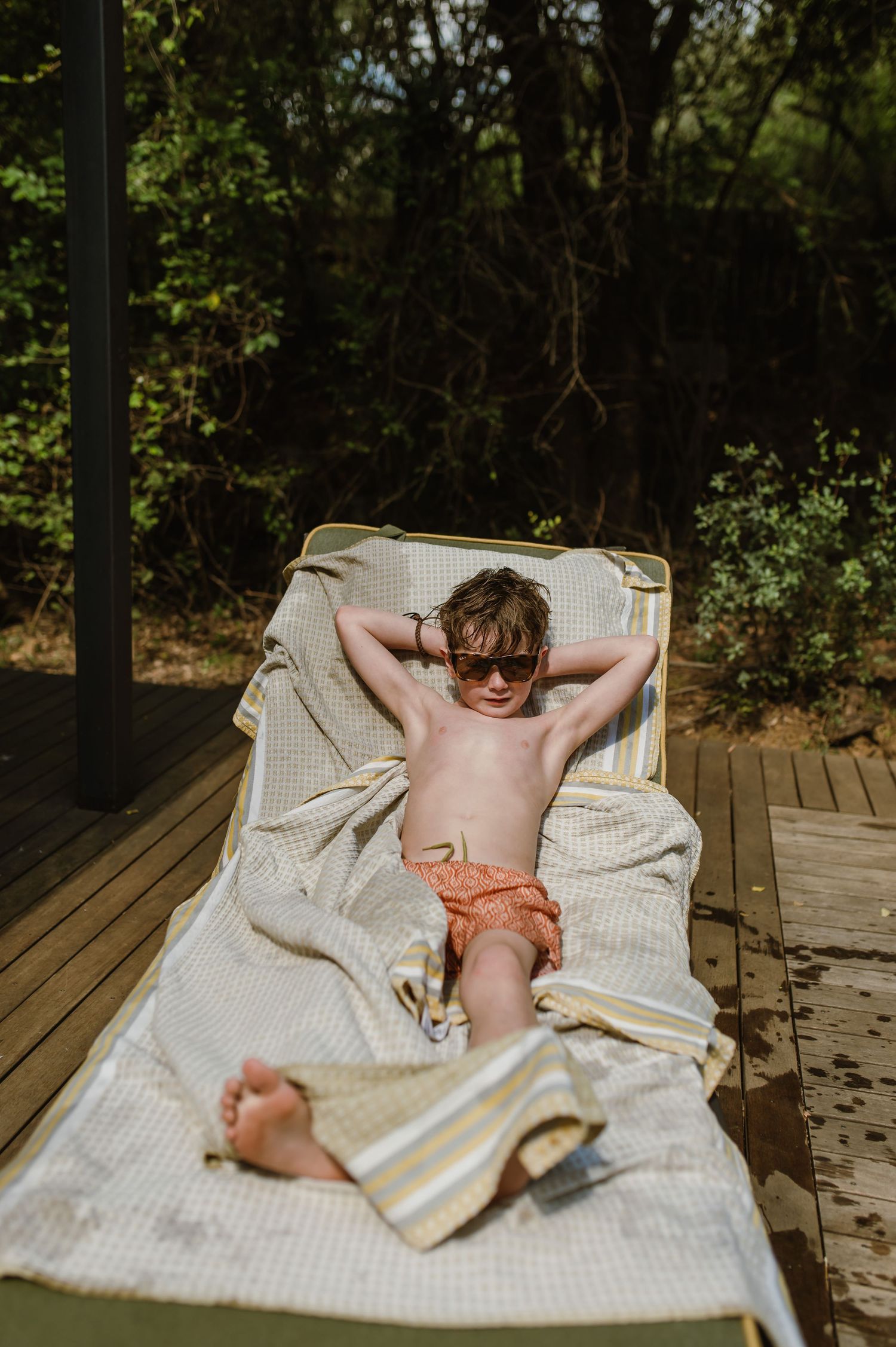 Young boy in sunglasses relaxing on a lounge chair with a yellow towel outdoors.