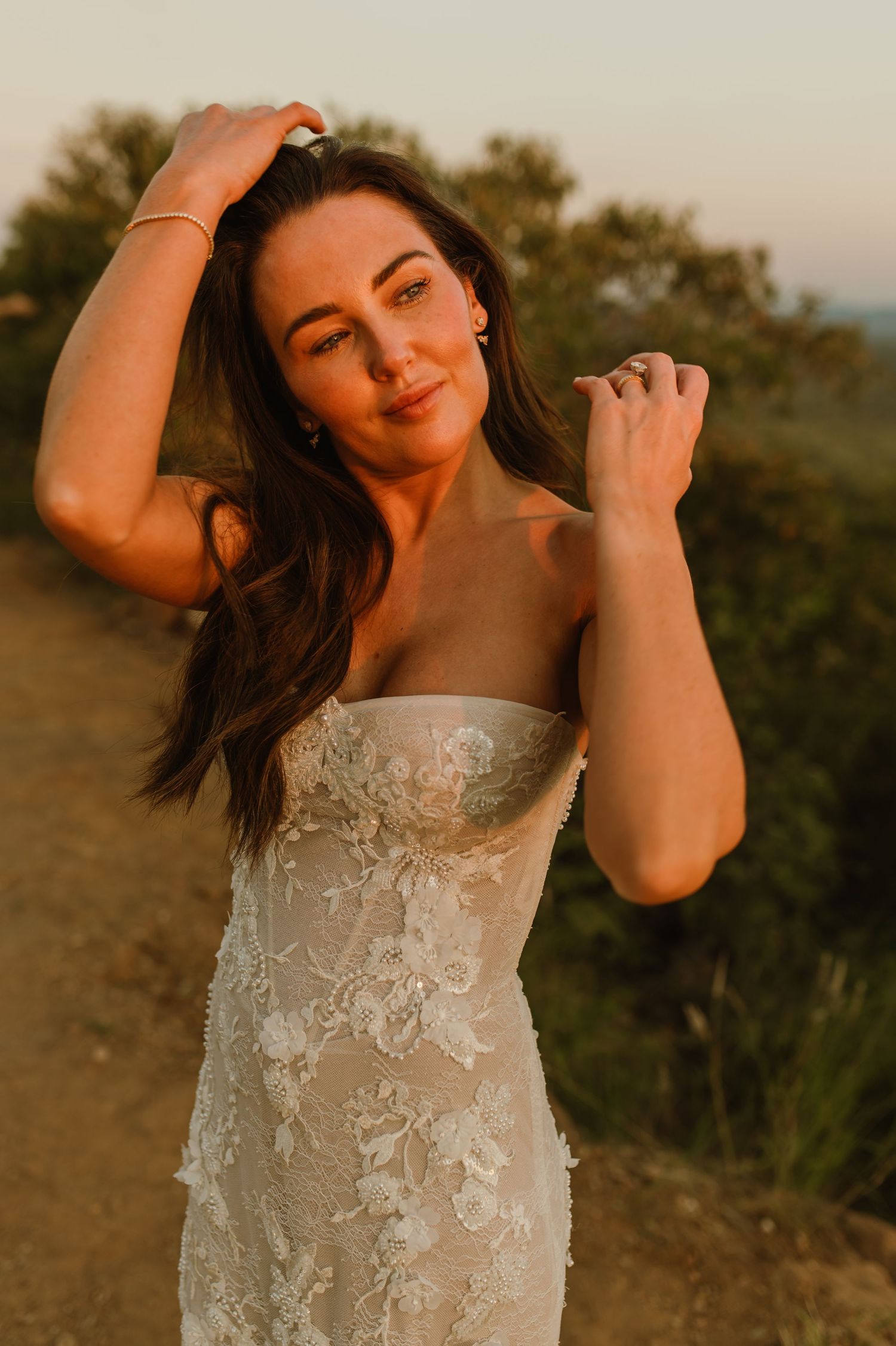 Bride in strapless floral lace wedding gown posing outdoors at golden hour sunset.
