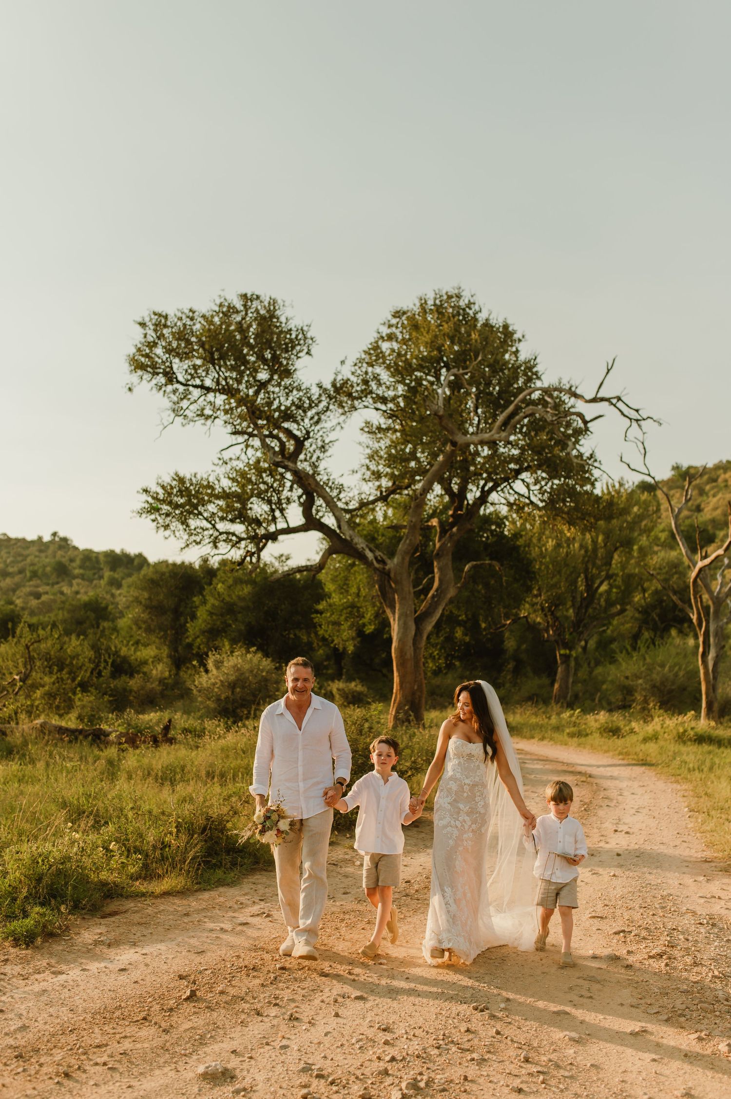 Family in white walks sandy path at golden hour with lush trees, bride in lace gown with veil, groom and two boys.
