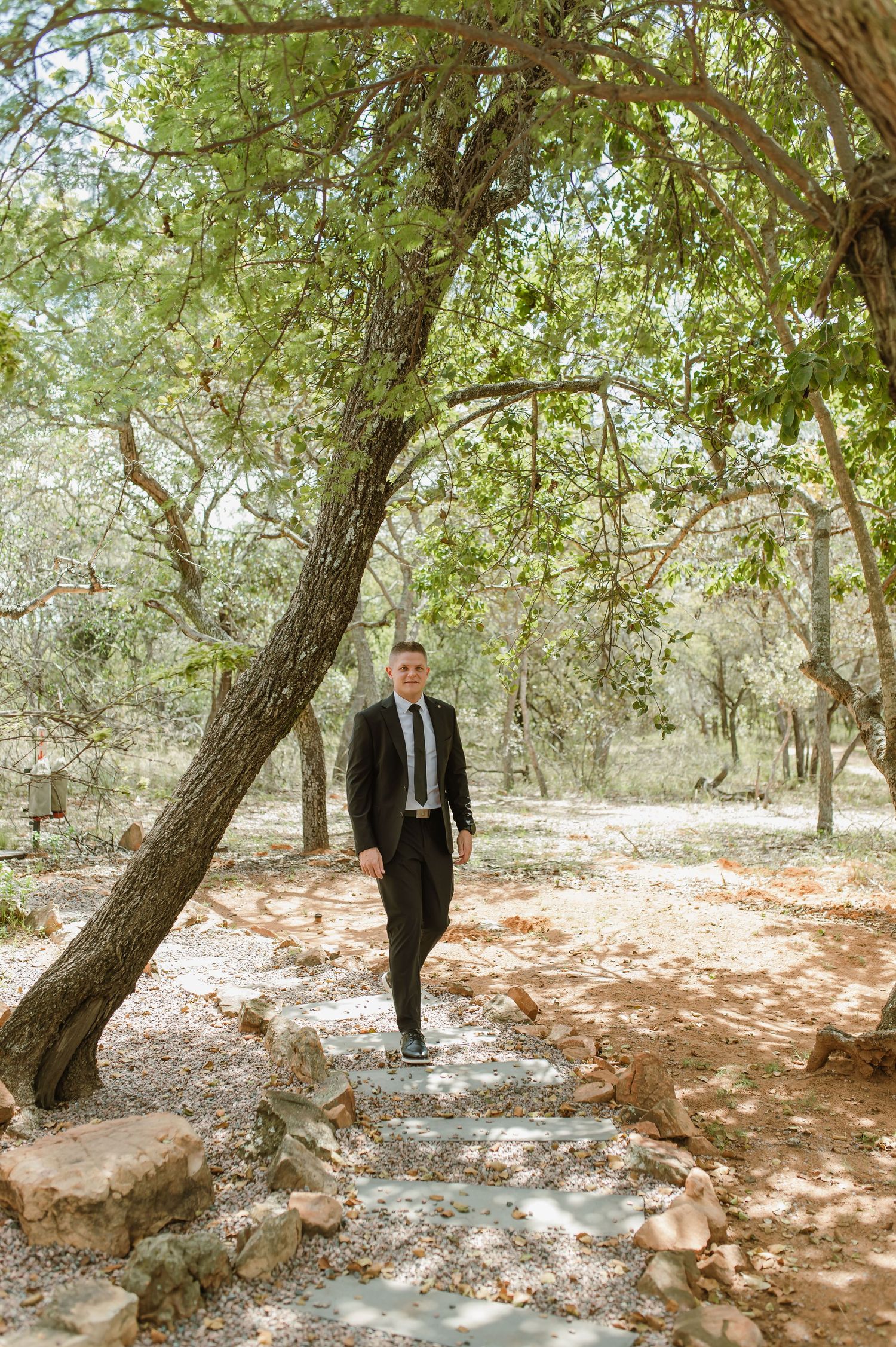 Dapper groom in black suit and tie walking along stone path through lush green woodland trees.