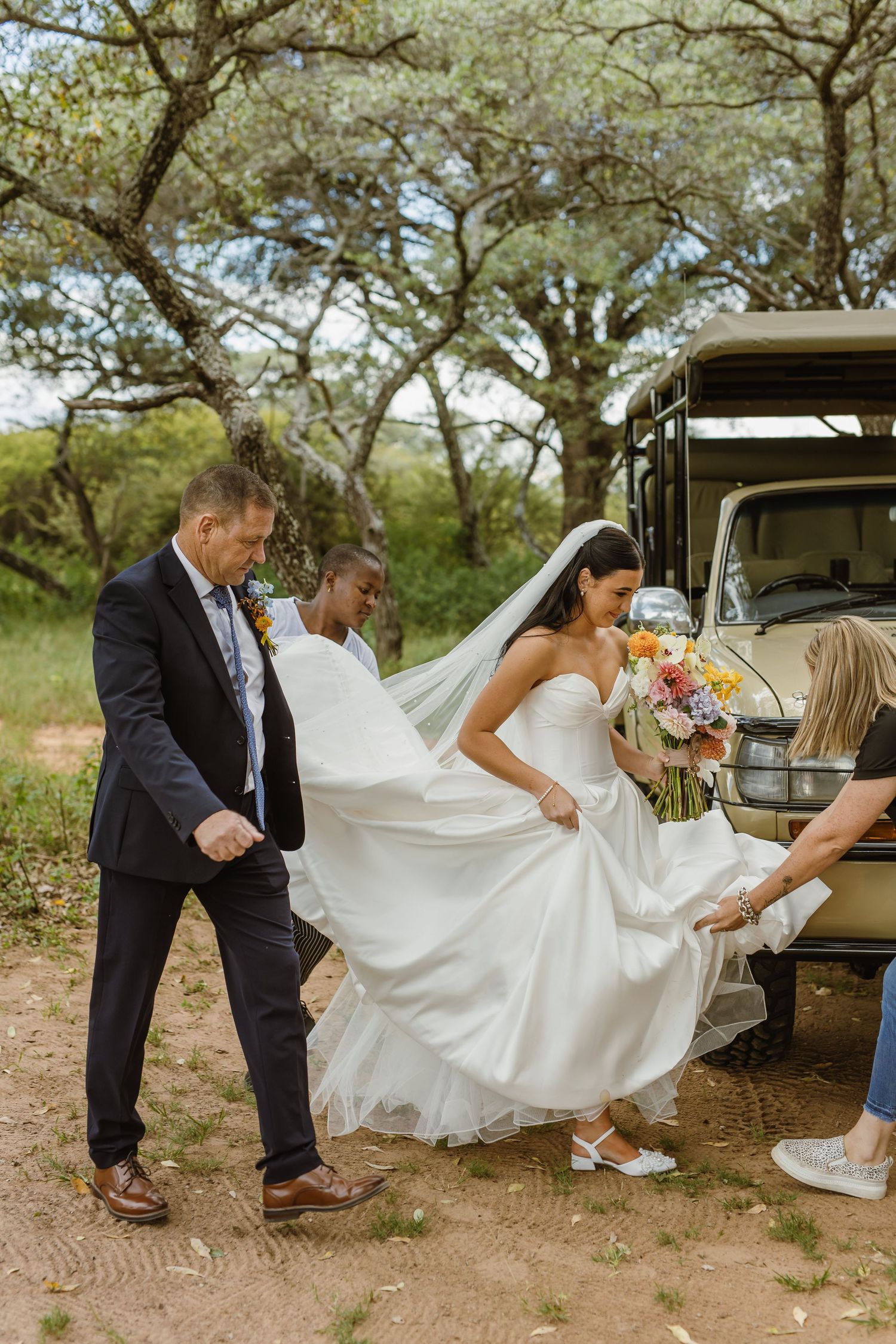 Bride in strapless gown with veil holding colorful bouquet being helped near vintage safari vehicle outdoors.