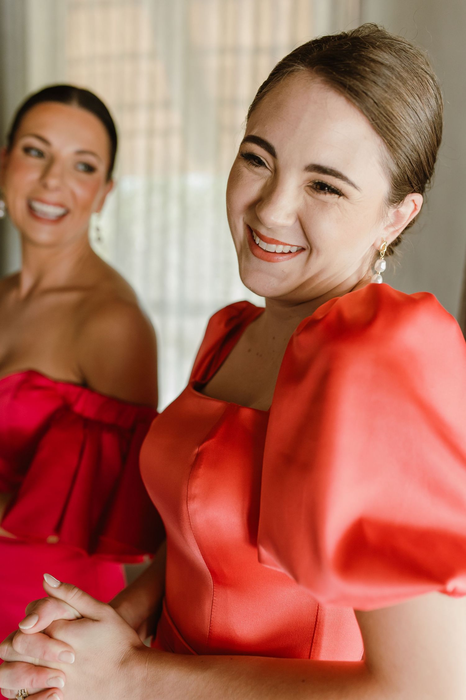Bridesmaids in red satin dresses laughing together while getting ready for wedding.