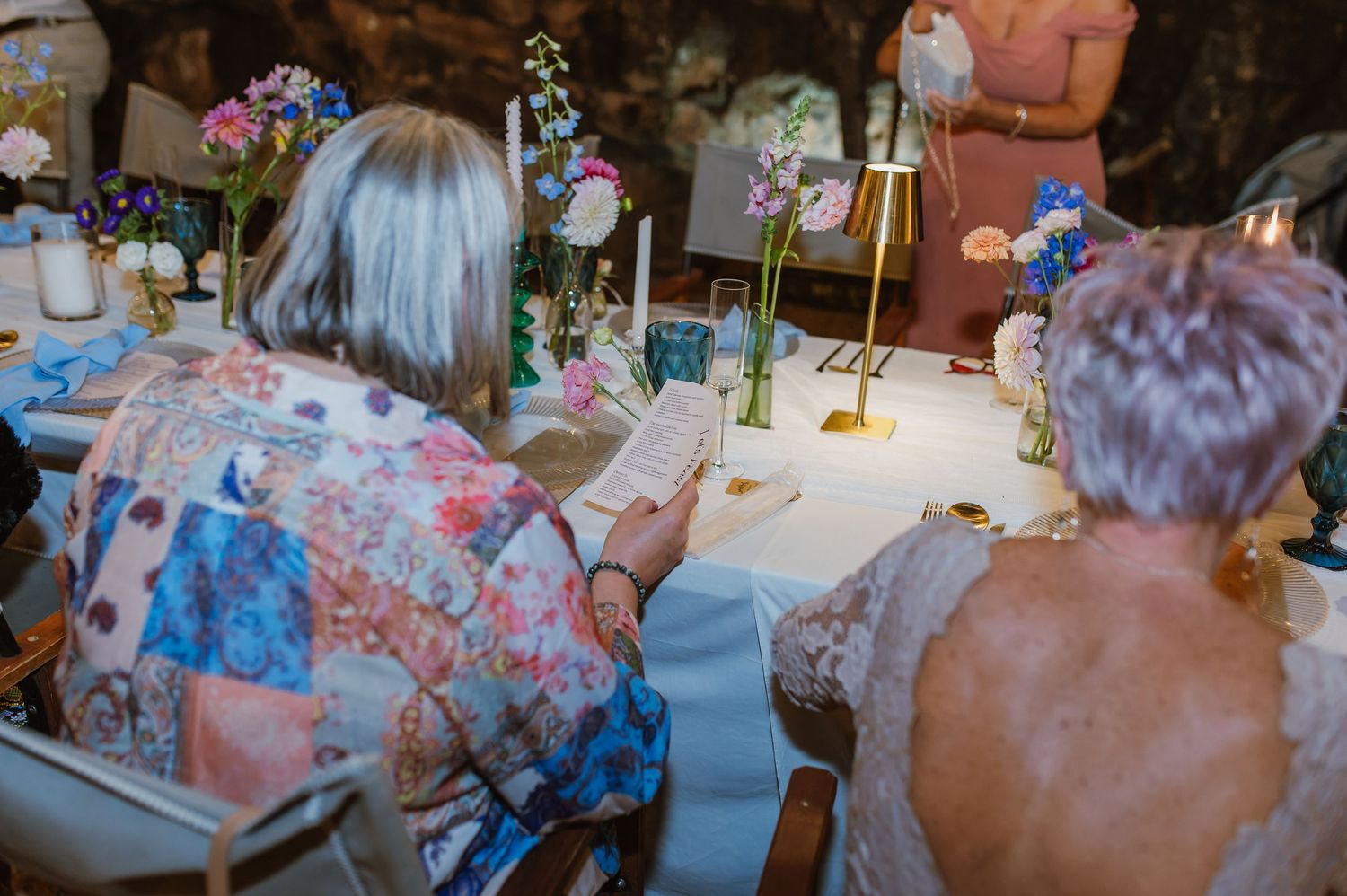 Wedding guests review programs at elegantly decorated reception table with colorful floral arrangements and candles.