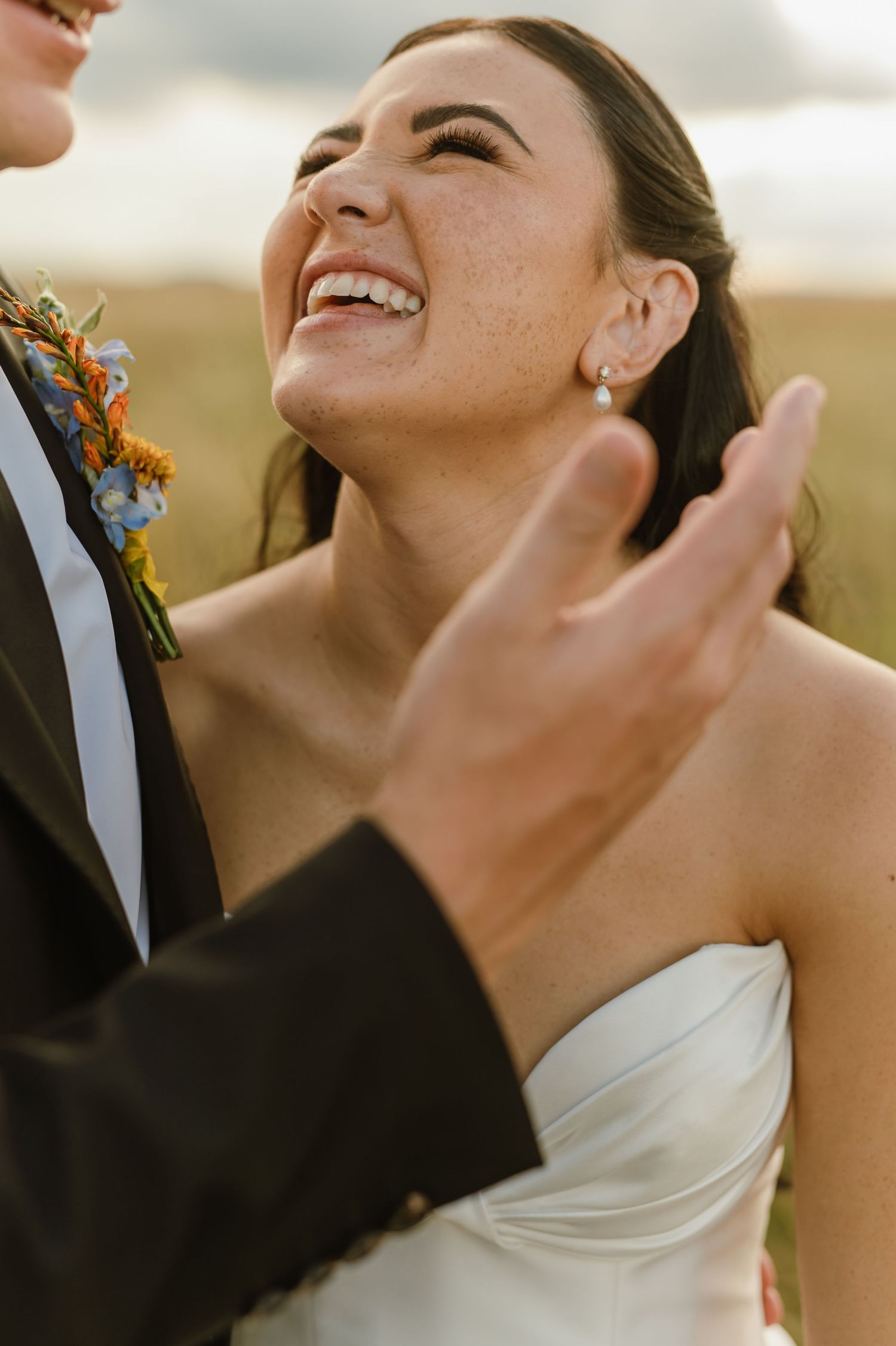 Joyful bride with freckles laughing in white strapless gown with pearl earrings during outdoor portraits.