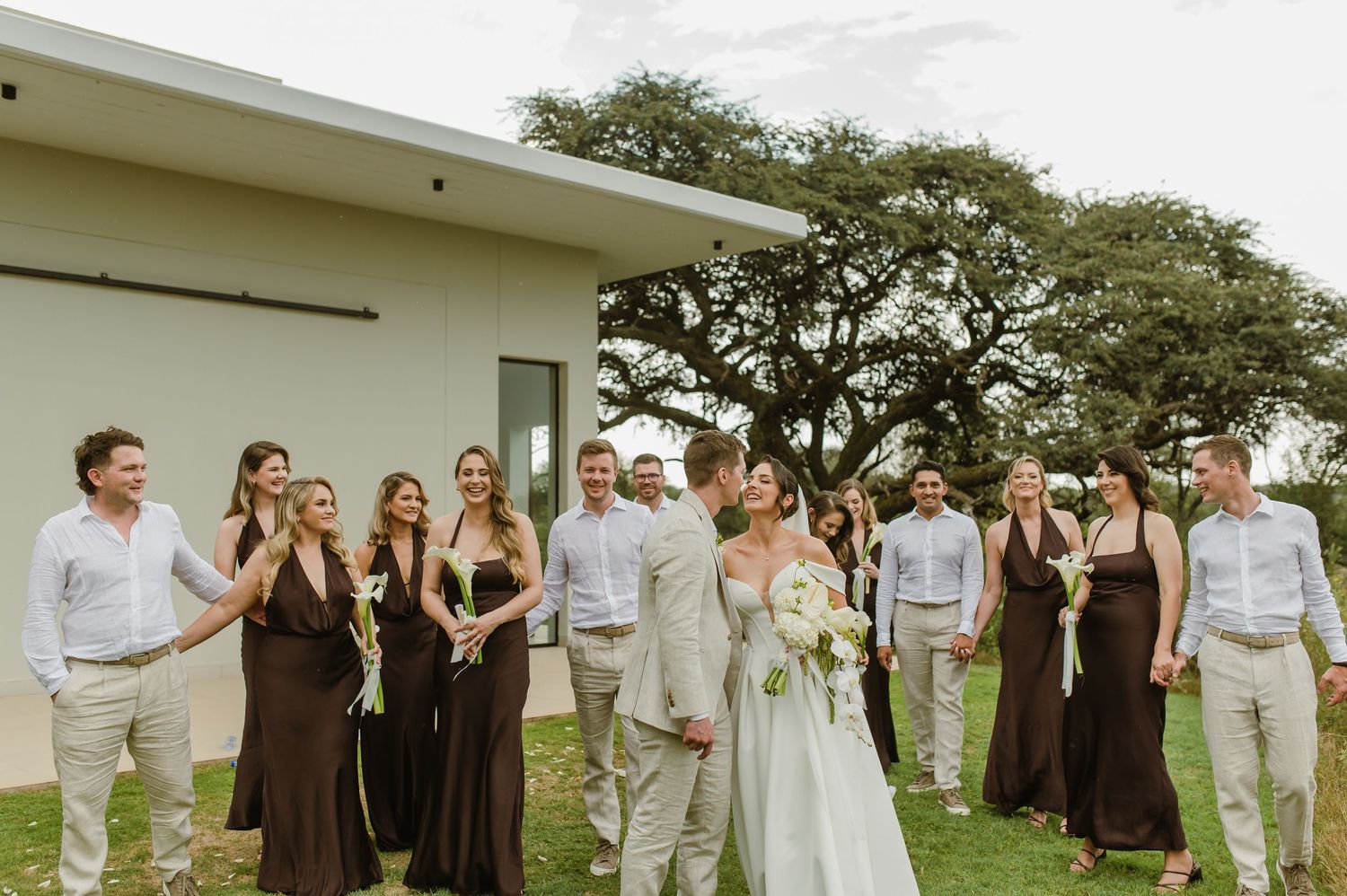 Wedding party in brown dresses and light colored suits stands together outdoors with trees in background.
