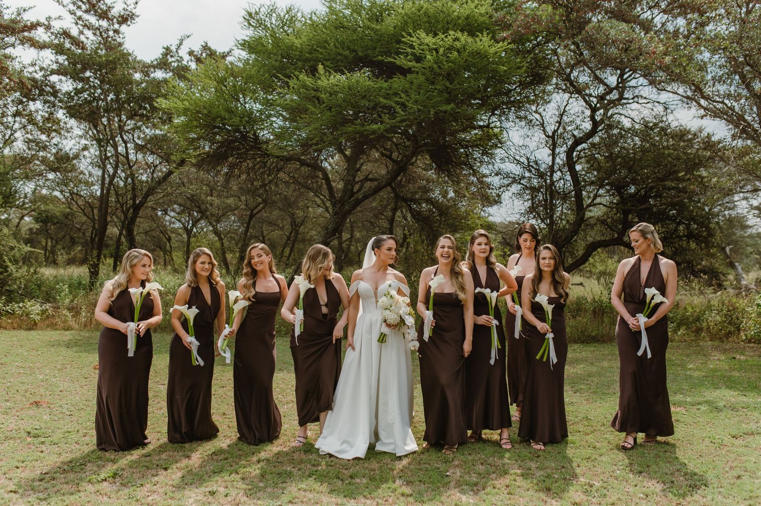 Wedding party in brown dresses poses together outdoors under green trees.