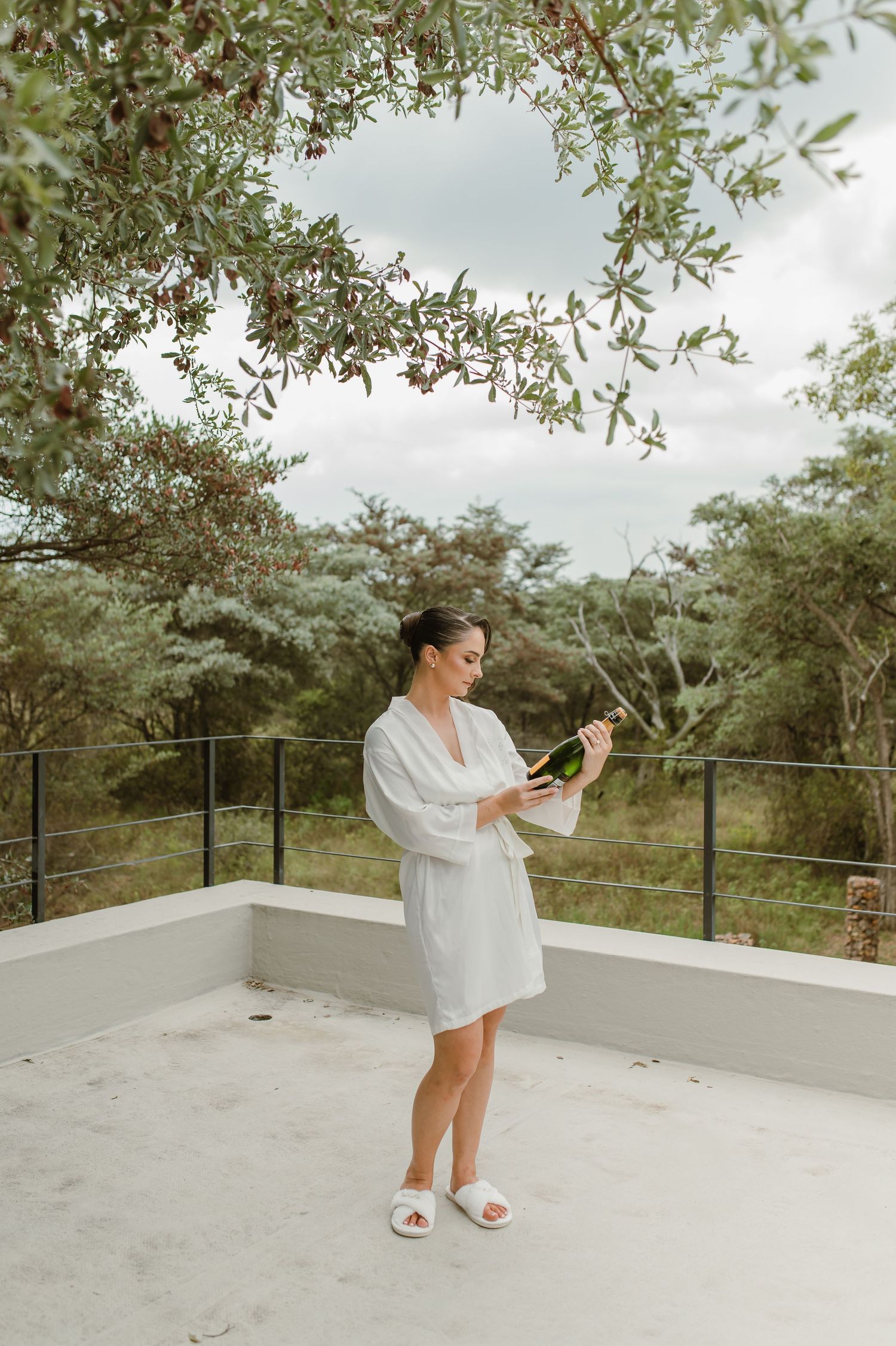 Person in white robe standing on modern balcony surrounded by olive trees.