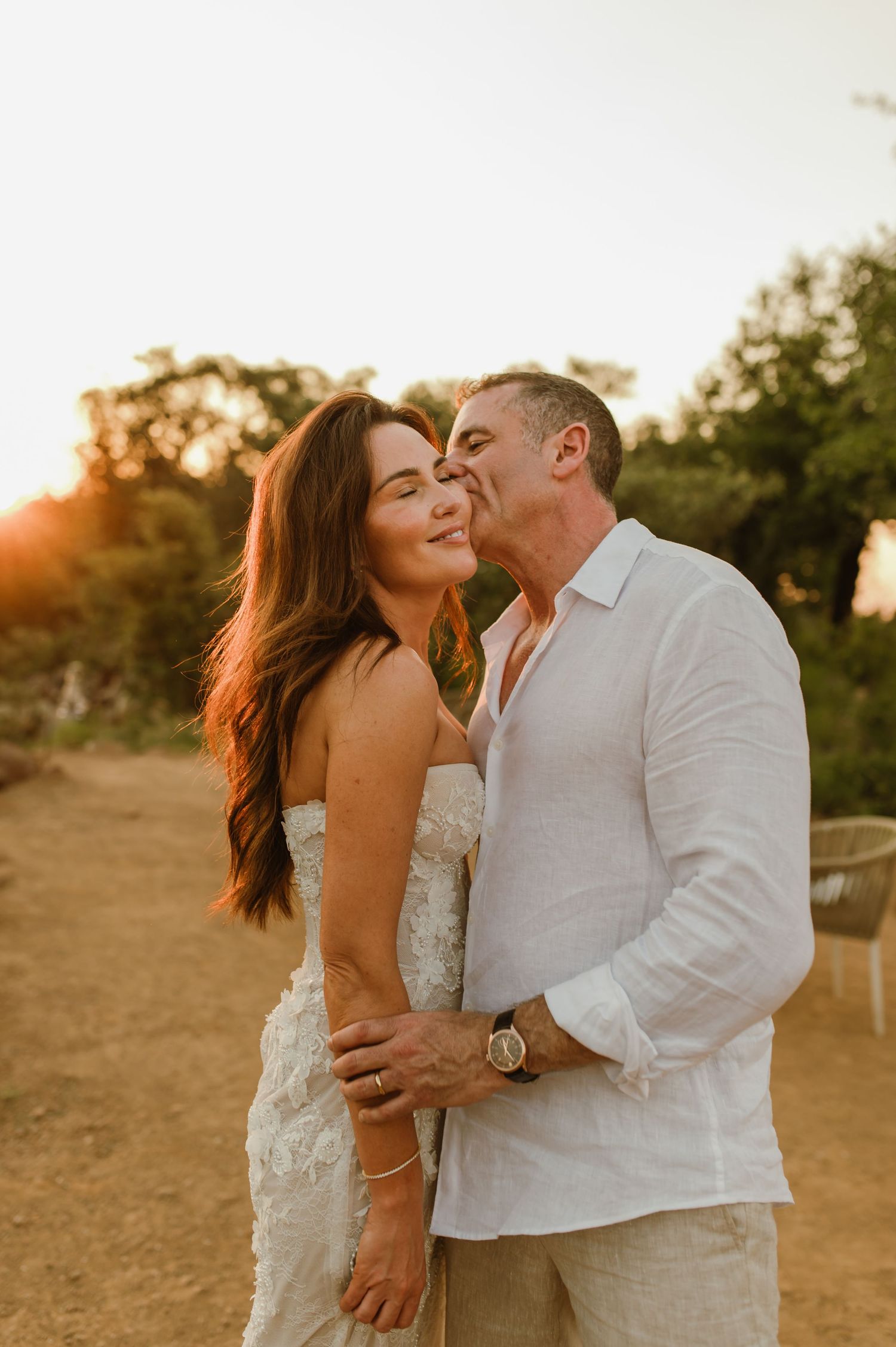 Groom kisses bride on cheek at sunset, she smiles in strapless lace wedding gown.