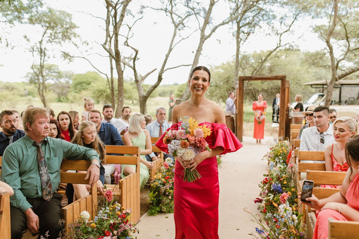 Bridesmaid in red off-shoulder dress walking down outdoor wedding aisle holding colorful bouquet.