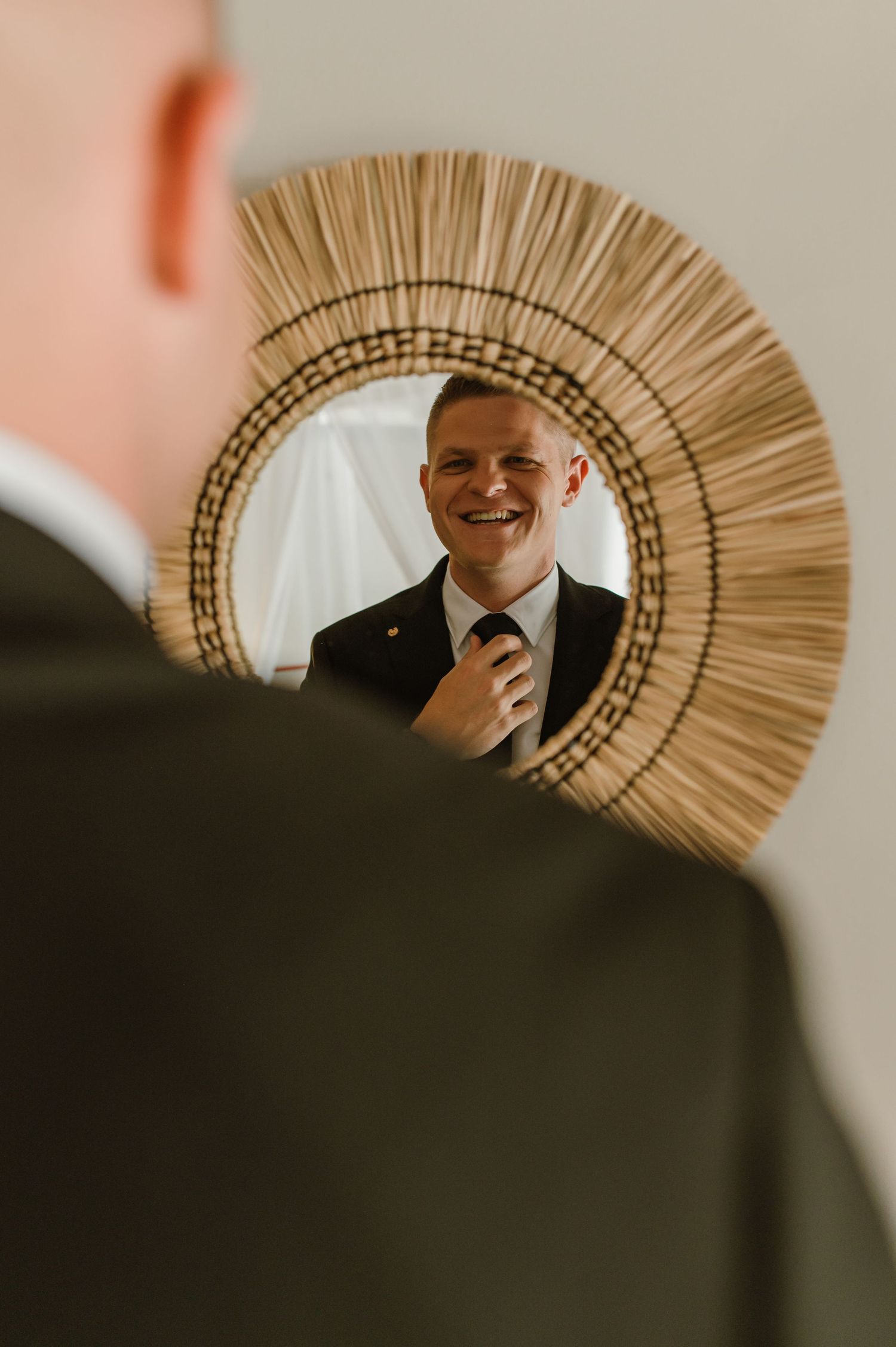 Smiling groom adjusting tie while looking at reflection in round woven rattan mirror before wedding ceremony.