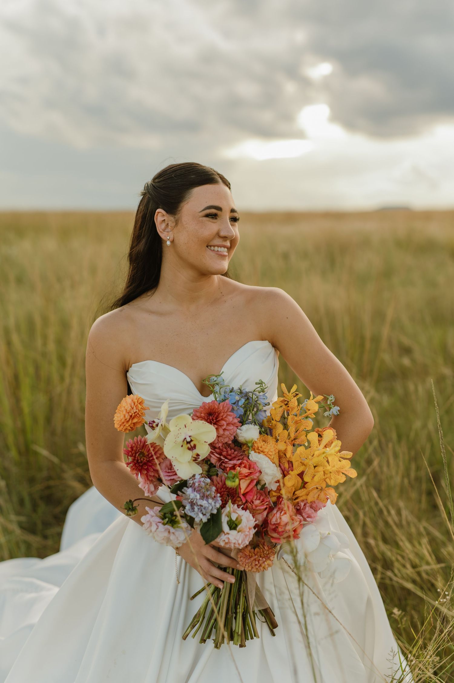 Radiant bride in strapless white gown holds colorful bouquet of dahlias and wildflowers in golden grass field.