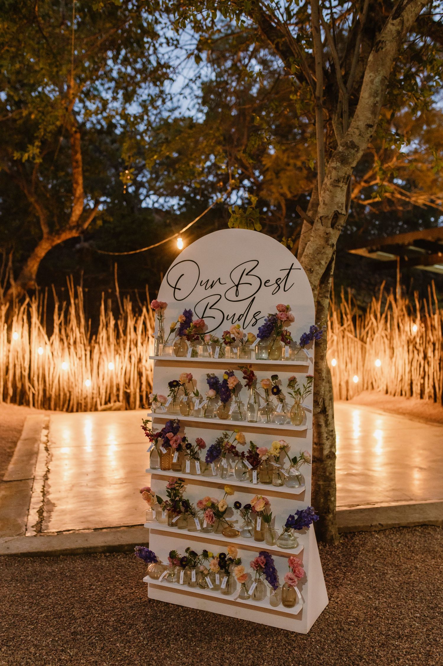 Elegant outdoor wedding display board reading Our Best Buds with colorful floral arrangements under string lights.