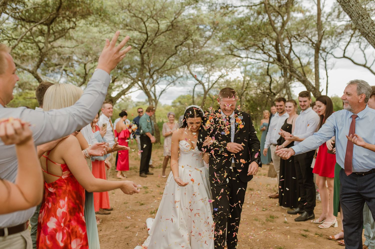 Newlyweds walking through a confetti shower as smiling wedding guests celebrate their outdoor ceremony exit.