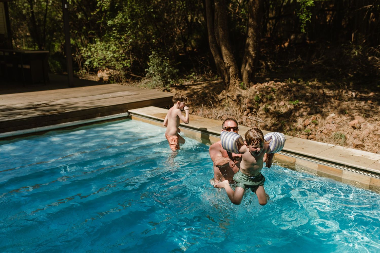 Children playing and jumping into a bright blue outdoor swimming pool surrounded by wooden decking and trees.