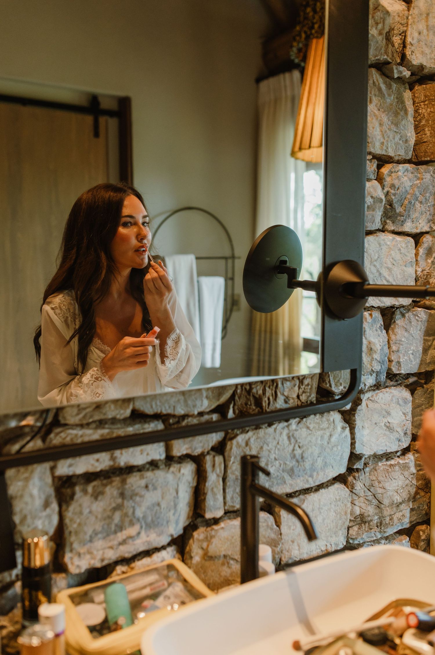 Woman applying lipstick in a rustic stone bathroom mirror surrounded by makeup and gold fixtures.
