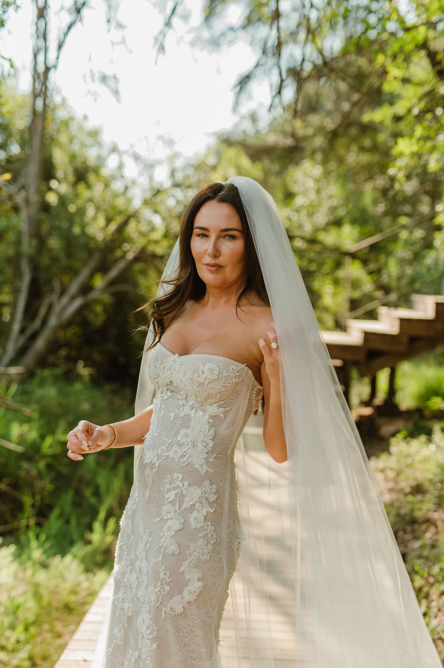 Bride in strapless lace wedding gown with flowing veil poses on outdoor wooden walkway.