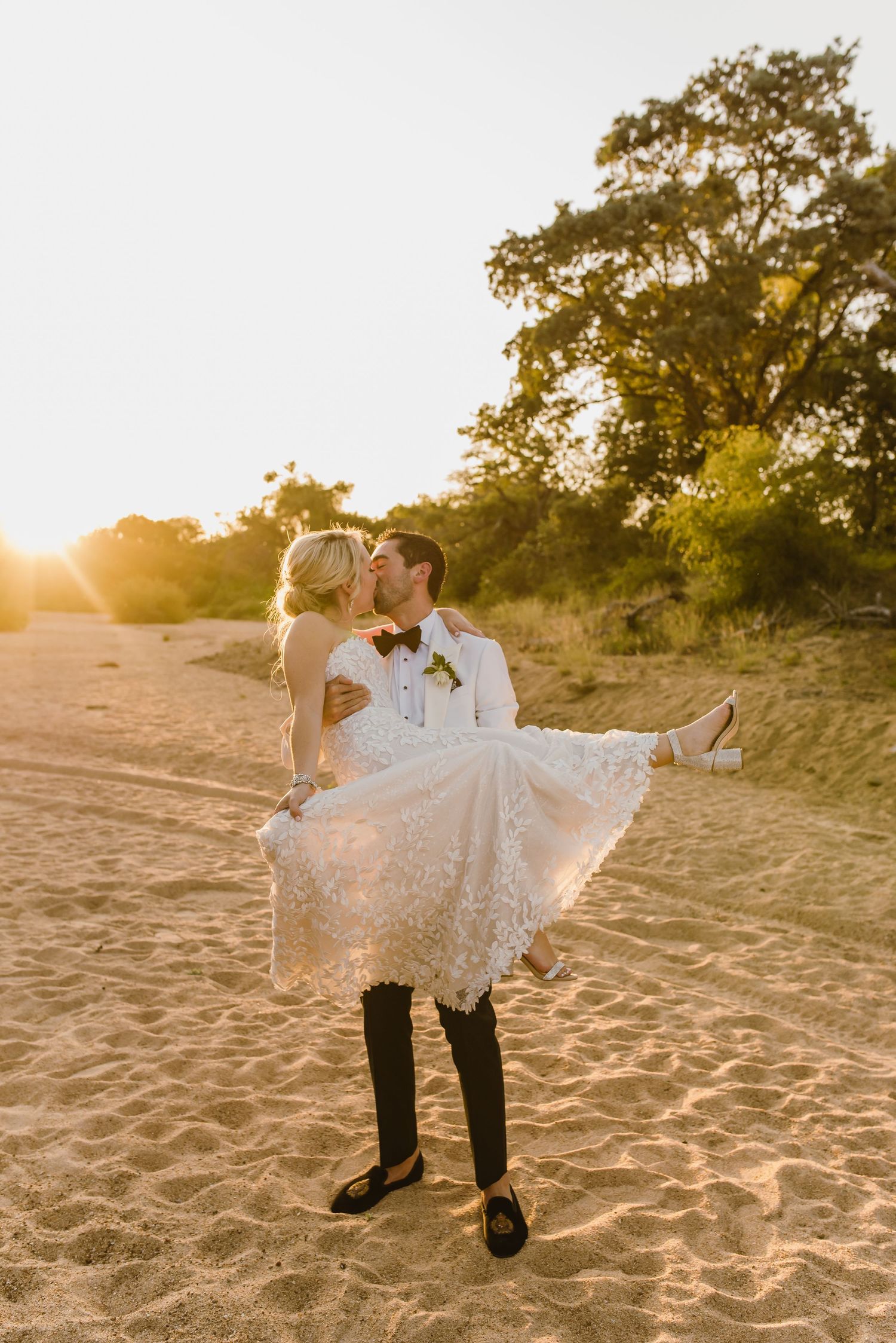 Romantic beach wedding photo with groom carrying bride during golden hour.